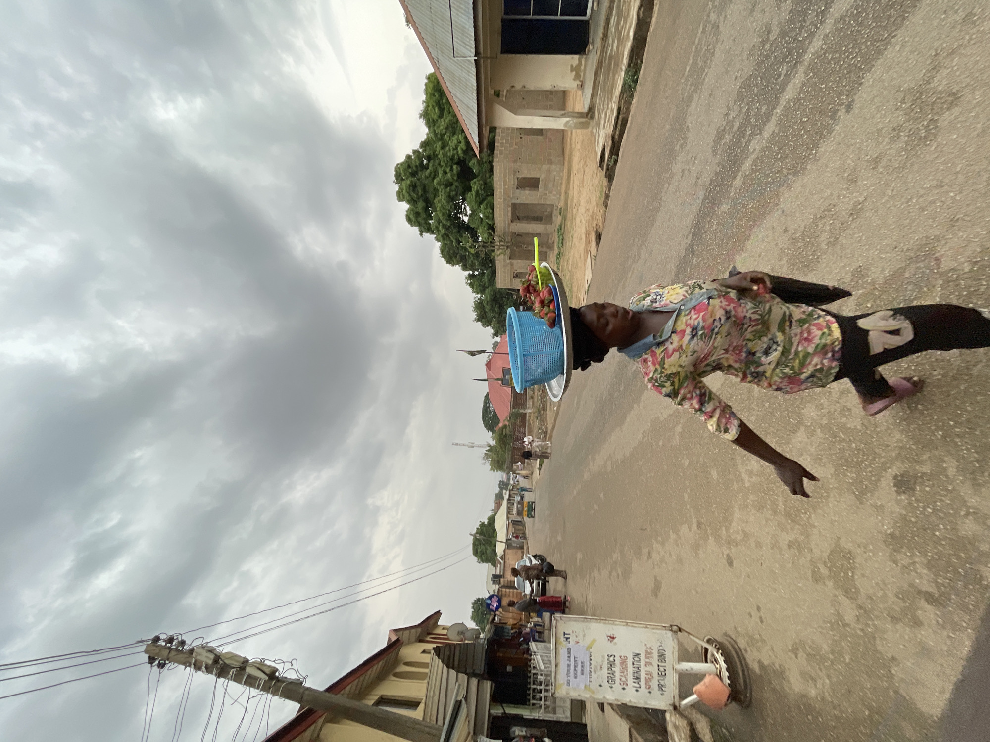 A woman walks down a street in Jos, Nigeria, balancing a tray on her head containing a blue basket with strawberries. She wears a floral shirt, black pants, and pink slippers. She extends one arm out to her side. A sign for graphic design and lamination services stands beside her on the right. Other buildings and people are visible in the background, along with a cloudy sky.