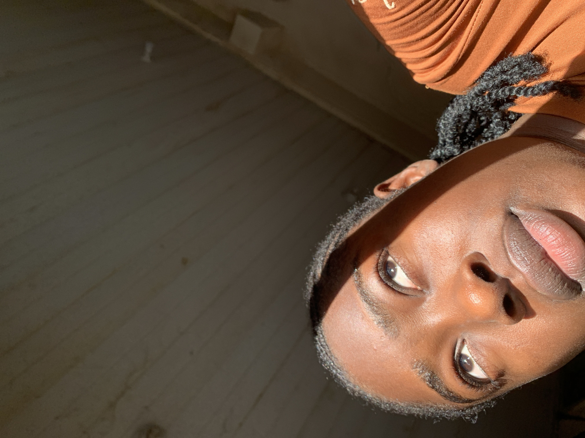 The scene depicts a close-up, angled portrait of a dark-skinned individual in an indoor setting within the City of New Stadium, Nigeria. The individual's face is positioned on the left side of the frame, with both eyes looking upwards and slightly to the right. A strong, warm light source from the upper left illuminates the left side of the face, casting a distinct, sharp diagonal shadow across the right forehead and face. Dark irises are visible in both eyes, and dark, well-defined eyebrows are present. The nose is partially visible in profile, and the full lips appear closed or slightly parted, exhibiting a natural, somewhat glossy texture. Dark, tightly coiled or braided hair is visible on the lower left, near the ear. A textured, orange-brown garment, likely a shirt, with visible folds, is present on the lower left. The background is dark and out of focus, appearing to be a wooden or paneled surface with diagonal lines, possibly a ceiling or upper wall. Faint lighter markings are discernible on this surface. A subtle, light-colored rectangular object, possibly an electrical fixture or outlet, is visible in the lower right background.