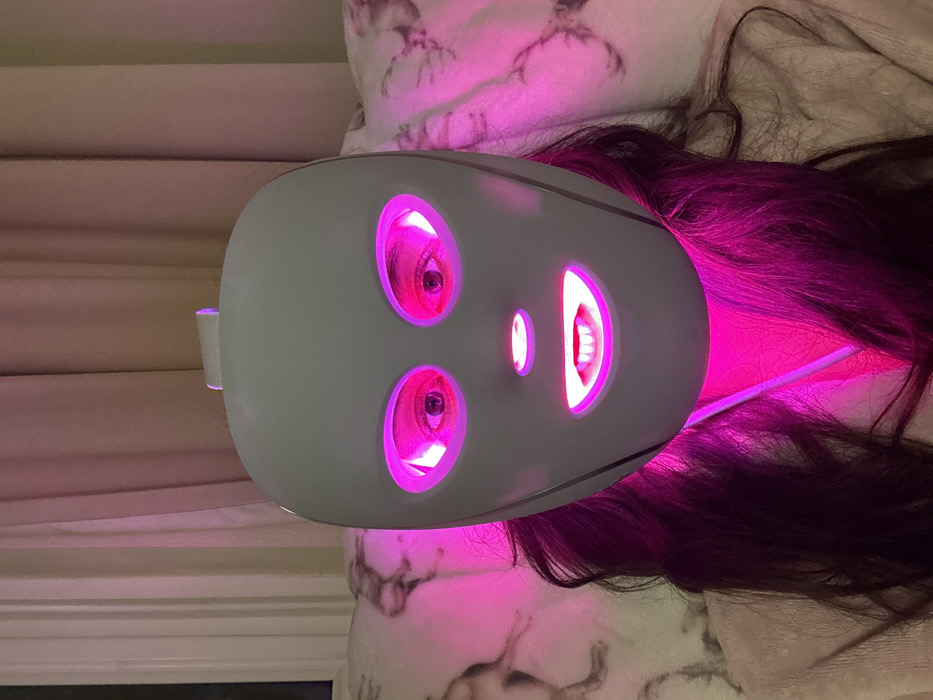 A person is wearing an LED face mask. The mask is gray and covers the eyes and mouth. Pink light is emitting from the eye and mouth openings. The person's eyes are visible through the eye holes. Their hair is dark and spread out around the mask. The person is lying on a patterned surface, possibly a pillow or blanket. A wall is partially visible in the background.