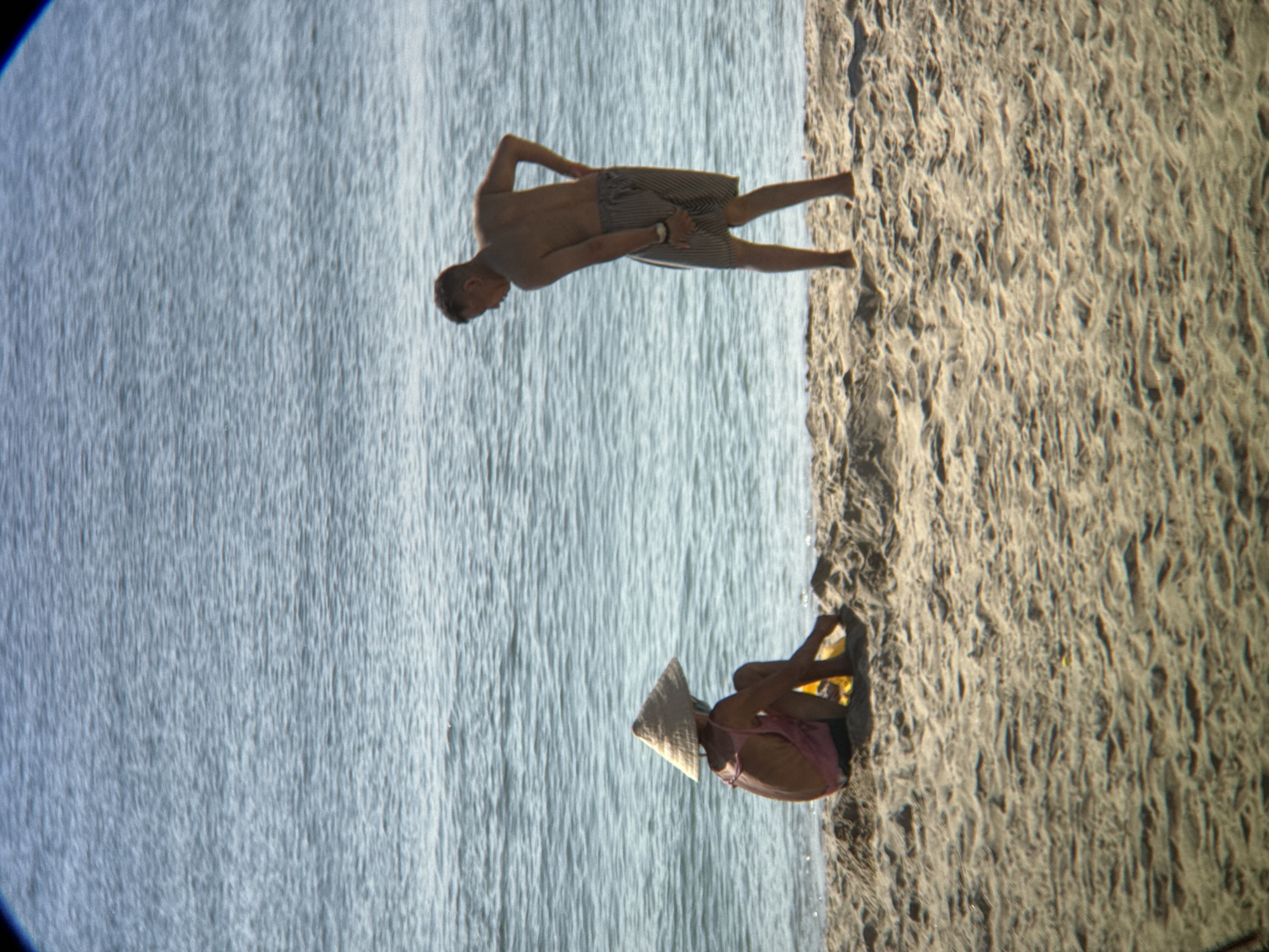 Two individuals are present on a sandy beach with the ocean in the background. The person on the left is wearing a conical hat and a pink garment and is sitting on the sand, facing away from the viewer. To the right, a shirtless person in striped shorts stands facing left, with their left hand on their hip and their right hand touching their shorts. The scene appears to be at a beach in Nha Trang, Vietnam, given the conical hat worn by one of the individuals, which is a traditional Vietnamese hat. The water is calm with gentle ripples, and the sand is light brown and textured with small mounds and indentations.