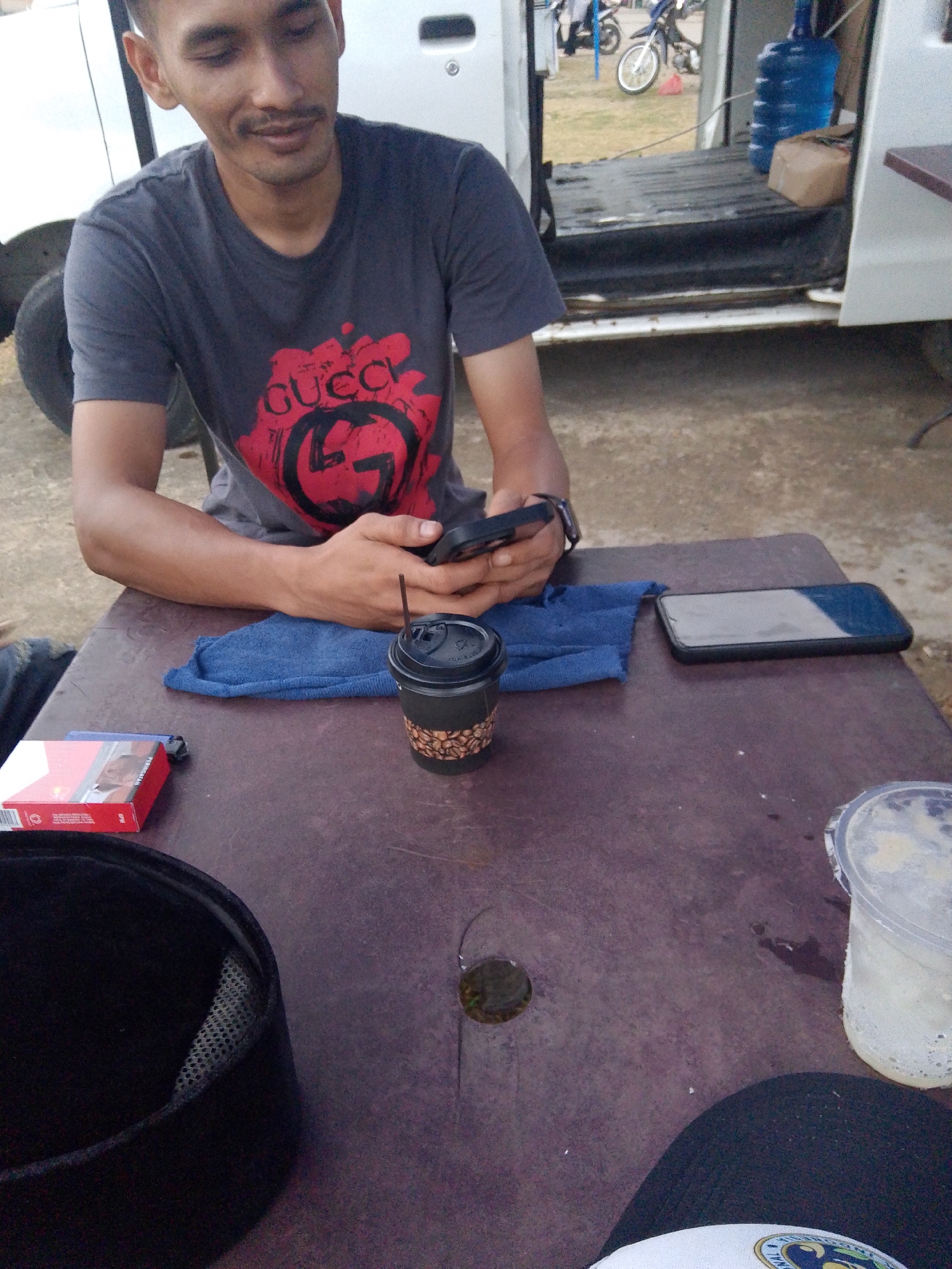 A young man with short dark hair and a mustache is sitting at a table, looking down at his phone. He is wearing a dark gray t-shirt with a red Gucci logo. On the table in front of him are a black coffee cup with a straw, a pack of red cigarettes, a blue cloth, and a smartphone. To the right of the table is a clear plastic cup with a white frothy drink. In the background, a white van is parked with its doors open. Outside the van, there are motorcycles and large blue water jugs. The ground is dirt and gravel.