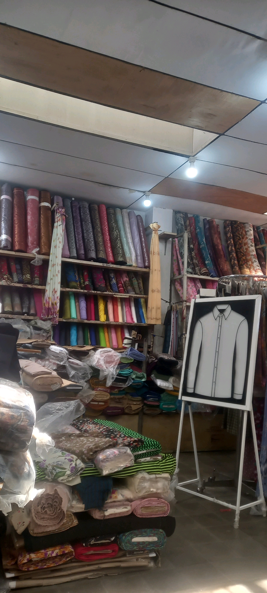 The image depicts the interior of a fabric shop in Jember, Indonesia. The shop is filled with shelves stacked high with various rolls of fabric, displaying a wide array of colors and patterns. Prominently displayed on an easel in the foreground is a framed illustration of a white collared shirt, serving as a display or advertisement. The ceiling is paneled with wooden and white sections, and illuminated by fluorescent lights. Rolls of fabric are also piled on the floor in the lower left section of the image, some covered in plastic. Clothing racks with hanging garments are visible in the background on the right. No individuals are clearly visible in the image.