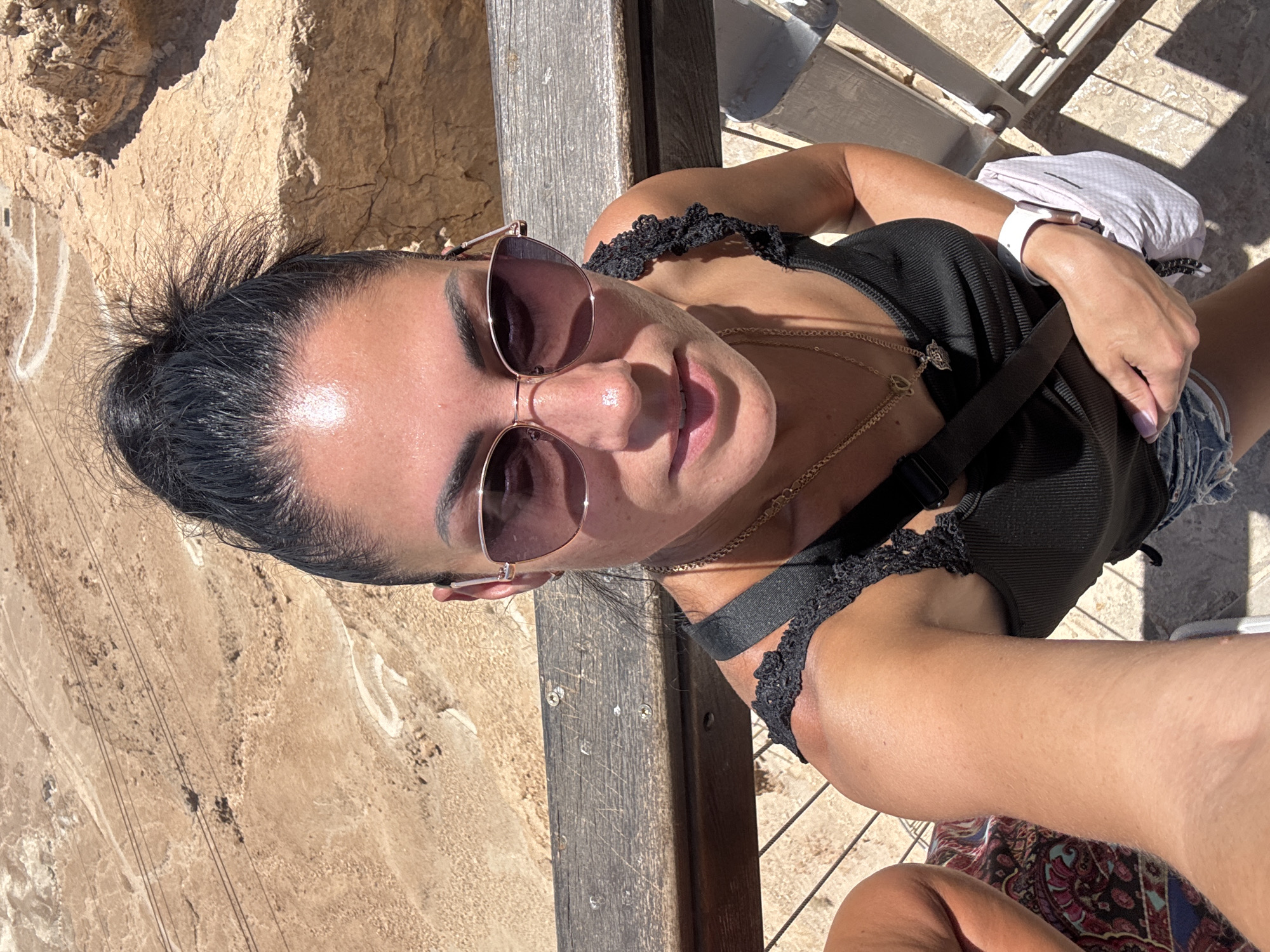 A woman with dark hair pulled into a high bun, wearing sunglasses and a black tank top with lace straps, takes a selfie. She is outdoors, standing on a wooden platform overlooking a rocky, desert landscape. A portion of another person's arm and leg are visible on the left. The woman is wearing necklaces and a smartwatch. A light-colored bag is slung across her body. The environment appears to be a tourist site in the Tamar Regional Council, Israel, likely Masada, given the distinctive arid terrain and ancient-looking rock formations.