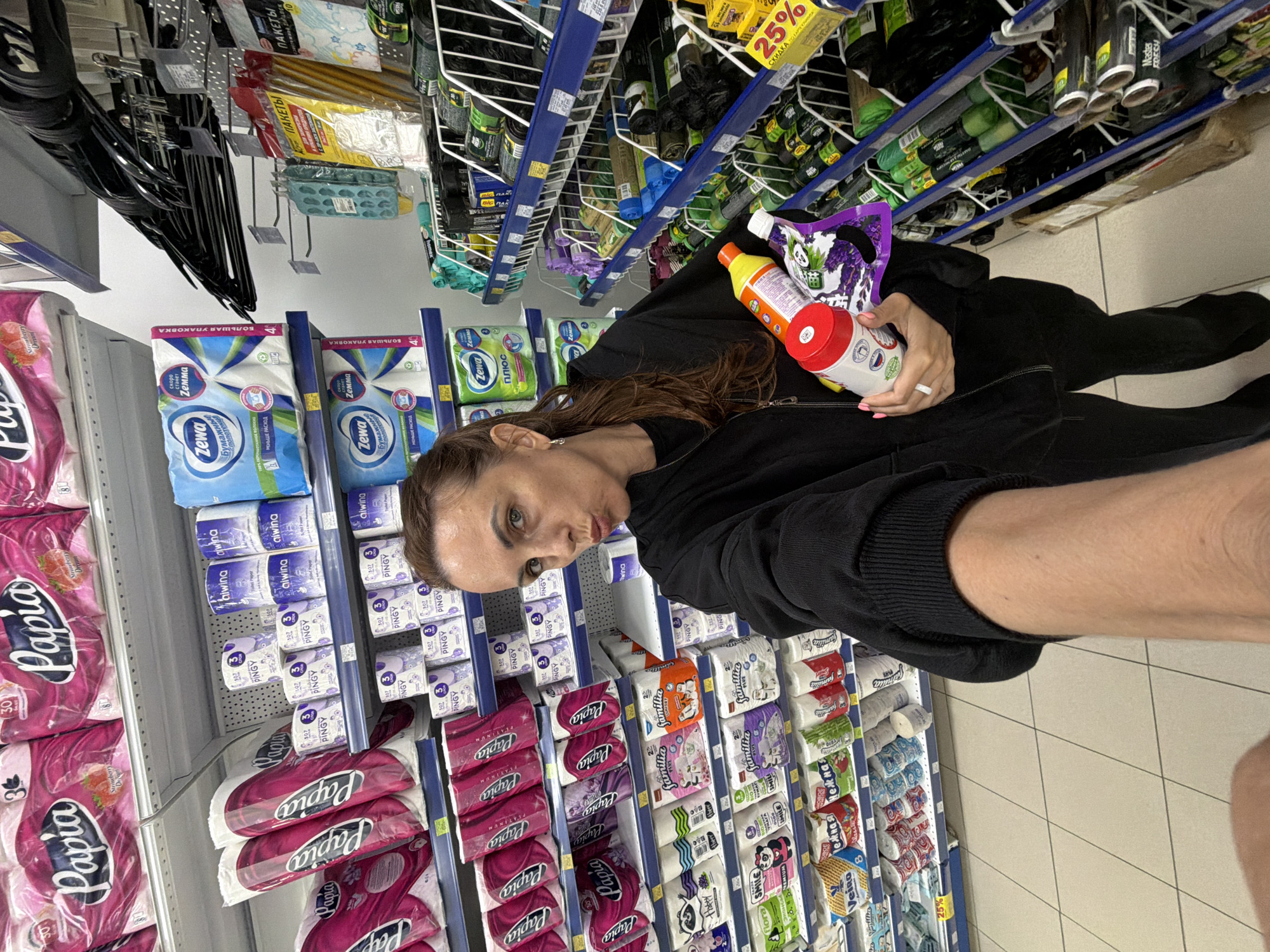 The image shows a woman in a store, likely a supermarket or hypermarket, standing in an aisle filled with paper products. She is holding two items, one is a red and white container, possibly a cleaning product or spray bottle, and the other is a purple packet with a panda illustration and lavender sprigs, which might be air freshener or a cleaning wipe package. The woman is looking towards the camera with a playful expression, making a puckered face. She is wearing a black hooded sweatshirt.
The shelves are stocked with various brands of paper towels and toilet paper, including "Papia," "Zewa," "alwina," and "PINGY." Some packages have clear branding, with one prominent display of "Papia" toilet paper in pink packaging and another featuring "Zewa" paper towels.
The background reveals more shelves with a variety of products, some of which appear to be cleaning supplies or household goods. Signs with text are visible, including one with "25%" and "СКИДКА ПО КАРТЕ КЛИВИТА" (25% discount with a Klievit card). Another sign reads "ПАКЕТЫ" (bags), and "Master FRESH". The store appears to be well-lit, typical of an indoor retail environment. The overall scene suggests a casual shopping trip.