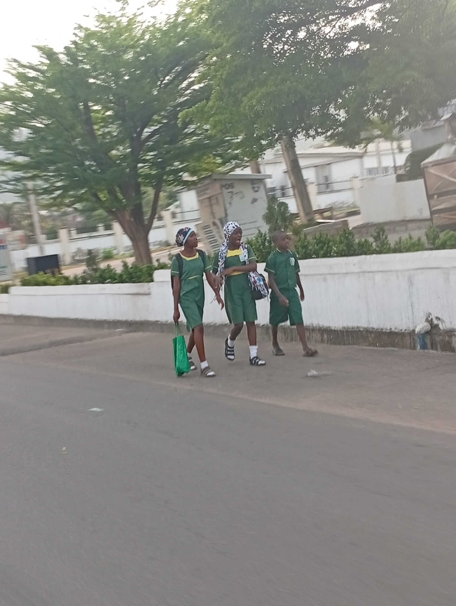 Three children walk along a paved road in Adankolo, Nigeria. They wear similar green school uniforms and head coverings. The children carry bags. A white wall runs behind them, separating the road from an area with trees and buildings.