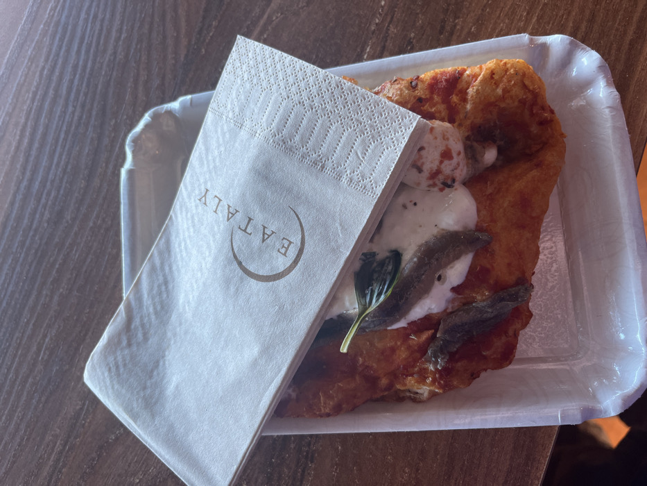 An overhead shot captures a single serving of an Italian dish, likely a fried pizza (pizza fritta), placed on a white, rectangular, disposable paper tray with slightly raised edges. The dish features a golden-brown, puffy, fried dough base, topped with white dollops of what appears to be mozzarella or burrata cheese, a few dark green basil leaves, and several silvery anchovy fillets.
A folded white paper napkin rests partially over the food on the left side of the tray. The napkin has a textured, patterned border along its top edge and prominently displays the word "EATALY" in a brown, sans-serif font, enclosed within a minimalist circular design.
The tray and its contents are situated on a dark, wood-grained surface, possibly a table or countertop. The lighting is even, suggesting an indoor setting, likely during the daytime. No people are visible, and the scene depicts a prepared meal ready for consumption in a casual dining environment, possibly within an Eataly food hall or similar establishment. The focus is entirely on the food and its presentation.