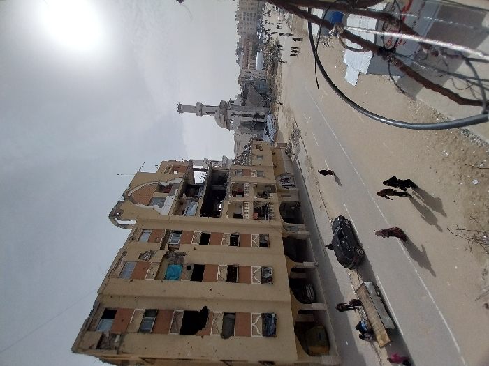 An elevated view captures a scene of extensive urban destruction in Khan Yunis, Palestinian Territory, under a bright, hazy sky.
In the foreground, the right side is partially obstructed by structural elements, including rebar and wires. A wide, paved road with visible lane markings extends from the foreground into the distance, flanked by dirt shoulders.
On the left, a large multi-story building, approximately six to seven floors high, shows significant structural damage. Large sections of its light-brown facade are missing, exposing interior rooms with broken windows and dark voids. The upper floors exhibit severe damage, including a partially collapsed arched architectural feature and exposed rebar. A yellow vehicle is partially visible within a ground-floor archway. Several individuals are gathered near the base of this building, with a group around a wooden cart being pulled by a person. A black sedan is parked in the left lane of the road.
Scattered pedestrians, including several women in traditional head coverings, walk along the road and its shoulders, casting long shadows.
In the mid-ground to background, a large mosque is visible, featuring a tall, slender minaret that appears mostly intact. The mosque's dome, however, shows substantial damage or partial collapse. Surrounding the mosque and further down the street, numerous other buildings are reduced to rubble or display varying degrees of destruction. In the far distance, more urban structures are visible, along with dispersed groups of people.