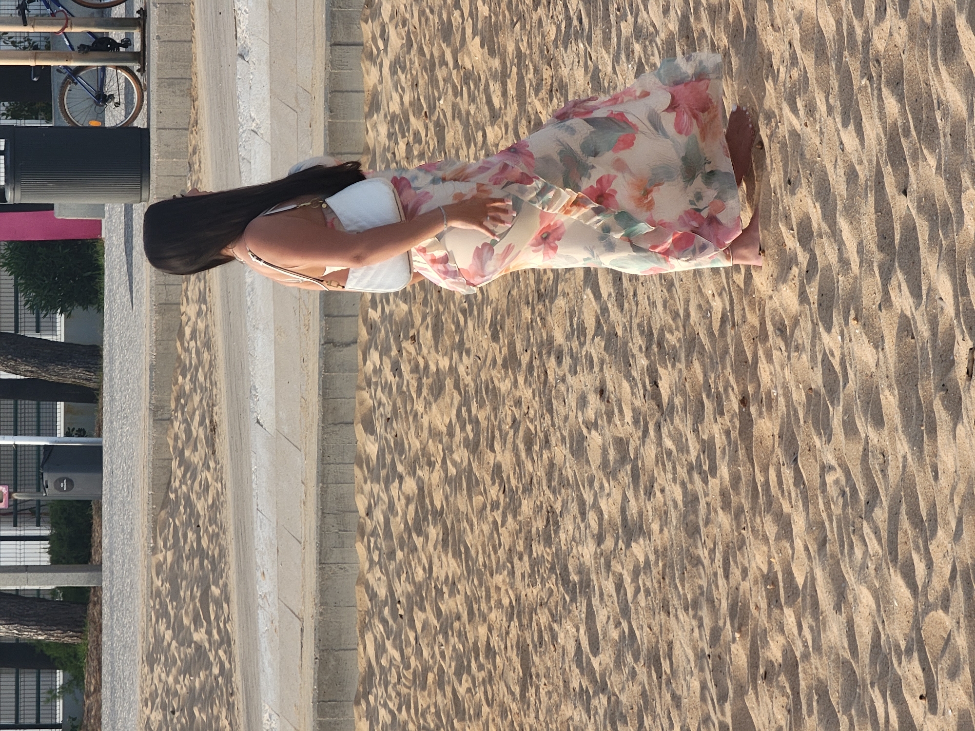 A young woman with long, dark hair stands on a sandy beach, facing right. She is wearing a flowing, floral-patterned maxi dress in shades of cream, pink, and green, with ruffles along the hem. She carries a white, crescent-shaped purse over her right shoulder. Her feet are bare and planted in the sand. Behind her, a paved walkway runs along the beach, separated by a low concrete barrier. In the background, a dark grey trash receptacle is visible, as is a bicycle with blue frame and brown wheels. The scene appears to be located in Quarteira, Portugal, a popular coastal town known for its beaches.