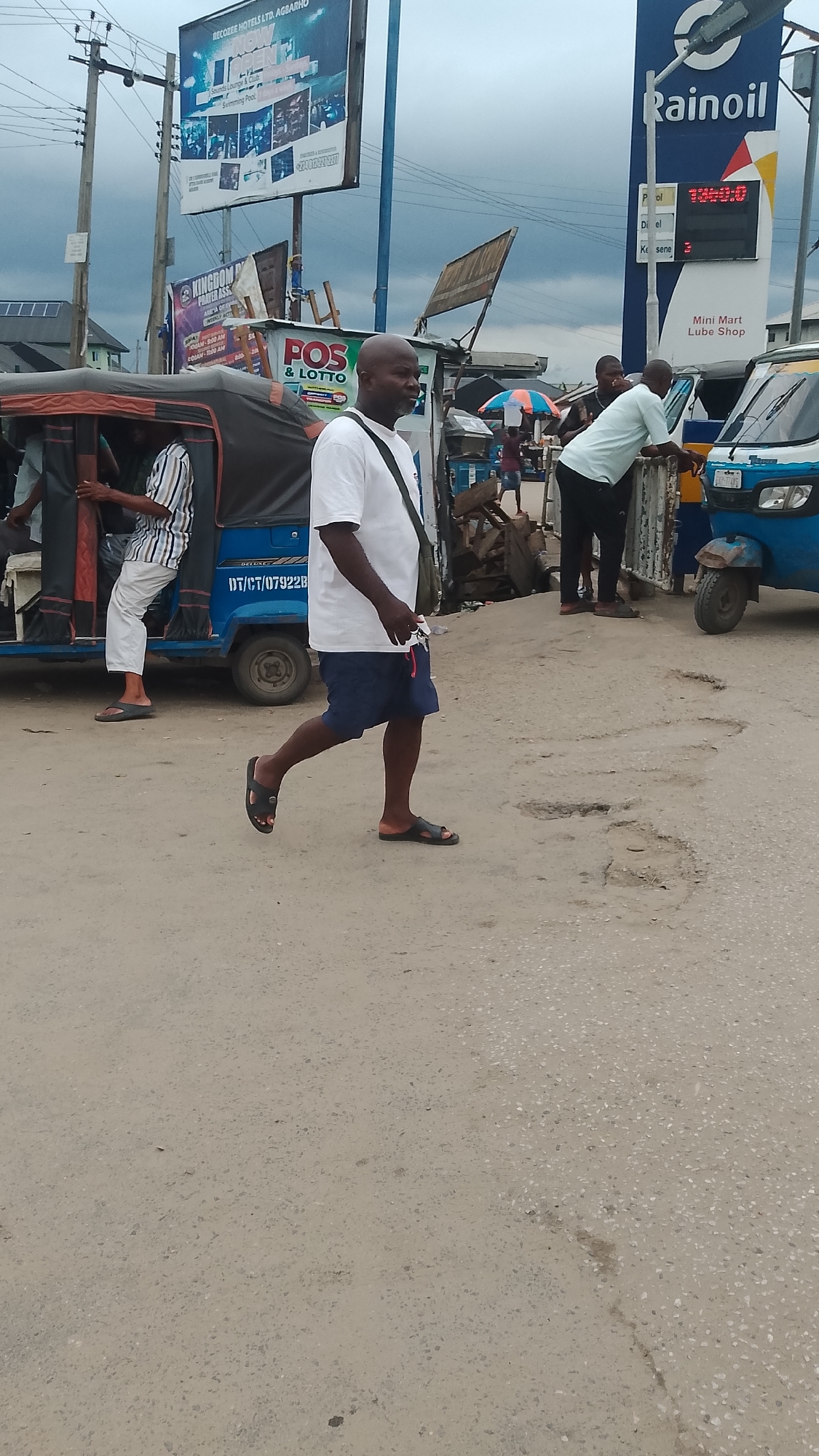 The image depicts a street scene in what appears to be Ukan, Nigeria, judging by the local signage. The street is paved with concrete, showing signs of wear and tear with potholes. Dominating the background are advertisements for "RECOZEE HOTELS LTD. AGBARHO," "KINGDOM PRAYER ASSK," and "POS & LOTTO," and a "Rainoil" petrol station with visible fuel prices. Several three-wheeled vehicles, presumably "keke napeps" or auto rickshaws, are stationed on the side of the road, with people entering or standing near them. A man is walking in the foreground, wearing a white t-shirt, blue shorts, and sandals. He has a shoulder bag. Another man is leaning against one of the rickshaws. The sky is overcast, suggesting possible rain.