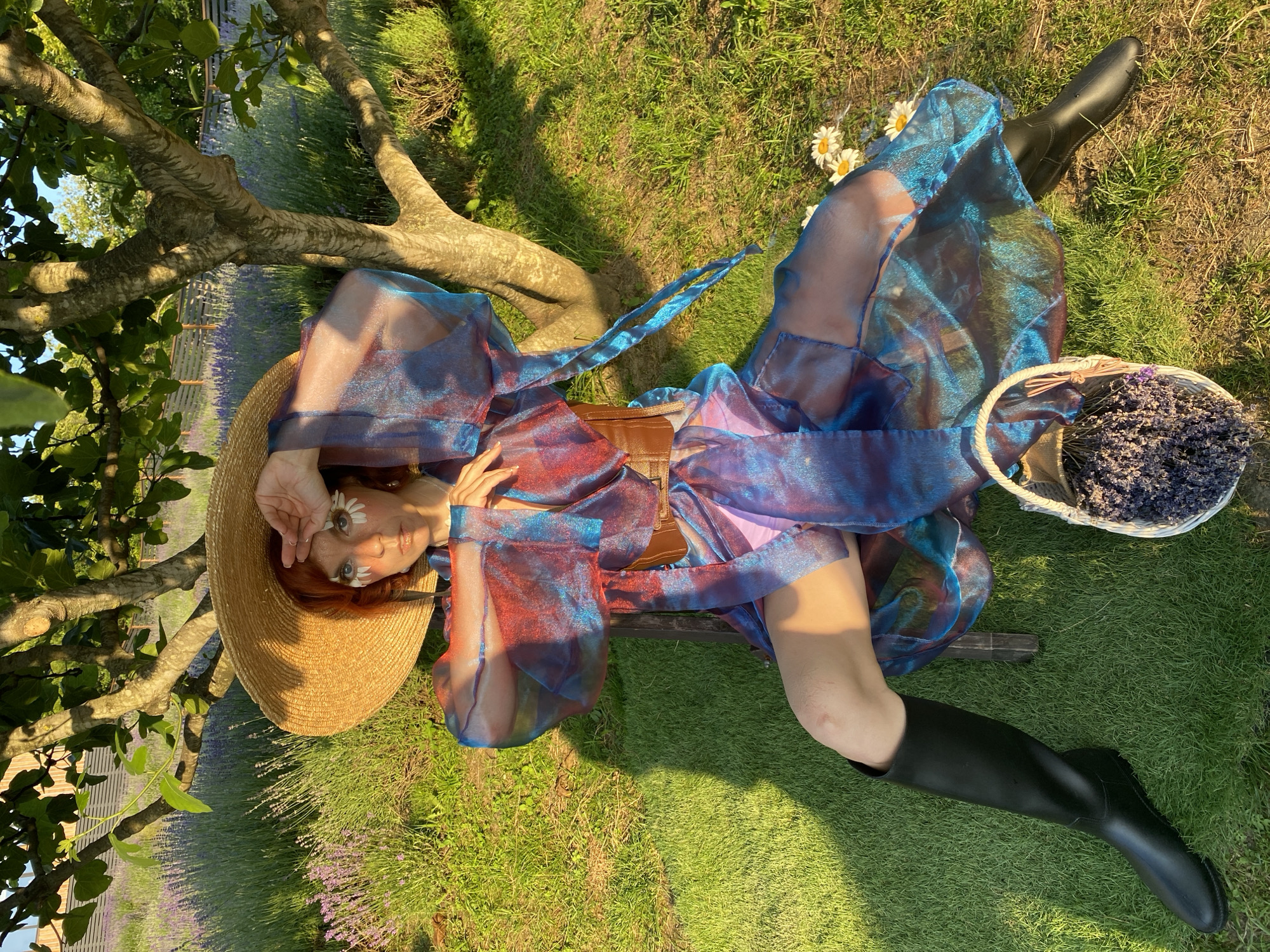A woman with bright red hair is lying on her back in a field of lavender, propped against the trunk of a tree. She is wearing a wide-brimmed straw hat, a shimmering blue and red dress, a brown corset, and black boots. Daisies are adorning her face. In her left hand, she holds a small basket filled with lavender. The sun is setting, casting long shadows across the grass. The location is Verkhneye Uchdere, Russia.