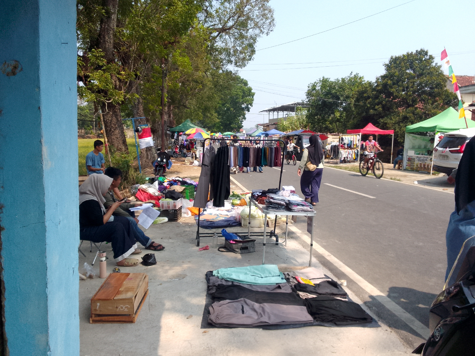 A street market is set up along a paved road in Mandalawangi, Indonesia. A variety of clothing is displayed on racks and laid out on the ground, suggesting a retail environment. Several people are present, some appearing to be vendors and others customers or passersby. One individual is seated on a chair, looking at a phone, while another is walking away from the camera. A person is riding a bicycle on the road. Tents and stalls are erected on the side of the road, some adorned with flags. The area is lined with trees, and the sky is clear and bright. The ground is a mix of concrete and dirt. A blue-painted structure is visible on the left side of the frame.