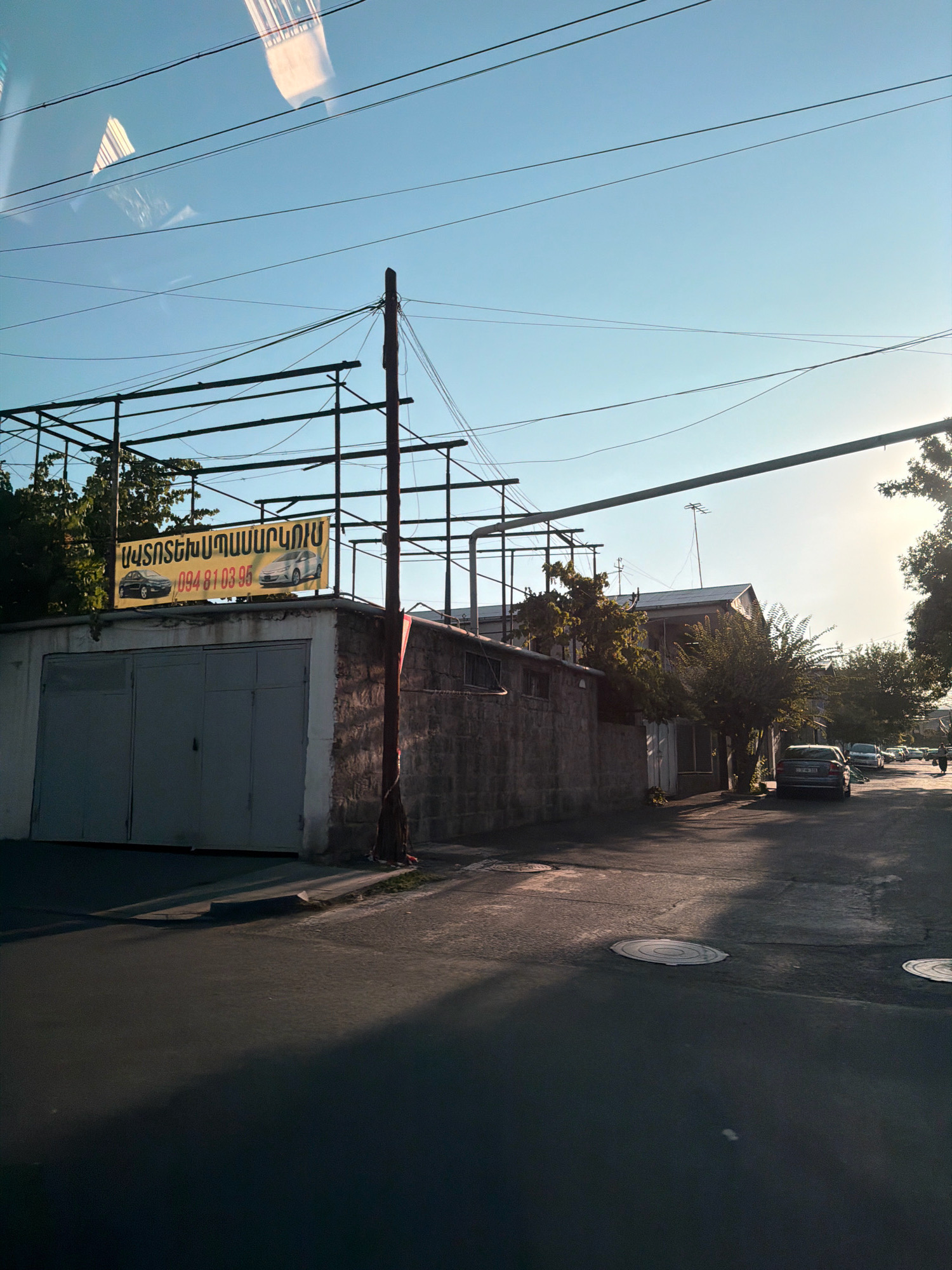 The image depicts a street scene in Yerevan, Armenia. In the foreground, the asphalt road is visible, with distinct shadows cast by sunlight. Several manhole covers are present on the road surface. To the left, a building with large gray garage doors is prominent. Above the garage doors, a yellow sign with Armenian text and images of two cars is displayed, along with a phone number. A dark-colored pole stands next to the building. The building itself appears to be constructed of stone blocks. Beyond this building, a street continues, lined with trees and other structures. Several parked cars are visible further down the street, and a person can be seen walking. Overhead, a network of power lines crisscrosses the sky, against a clear blue sky. The lighting suggests it is late afternoon or early evening.
