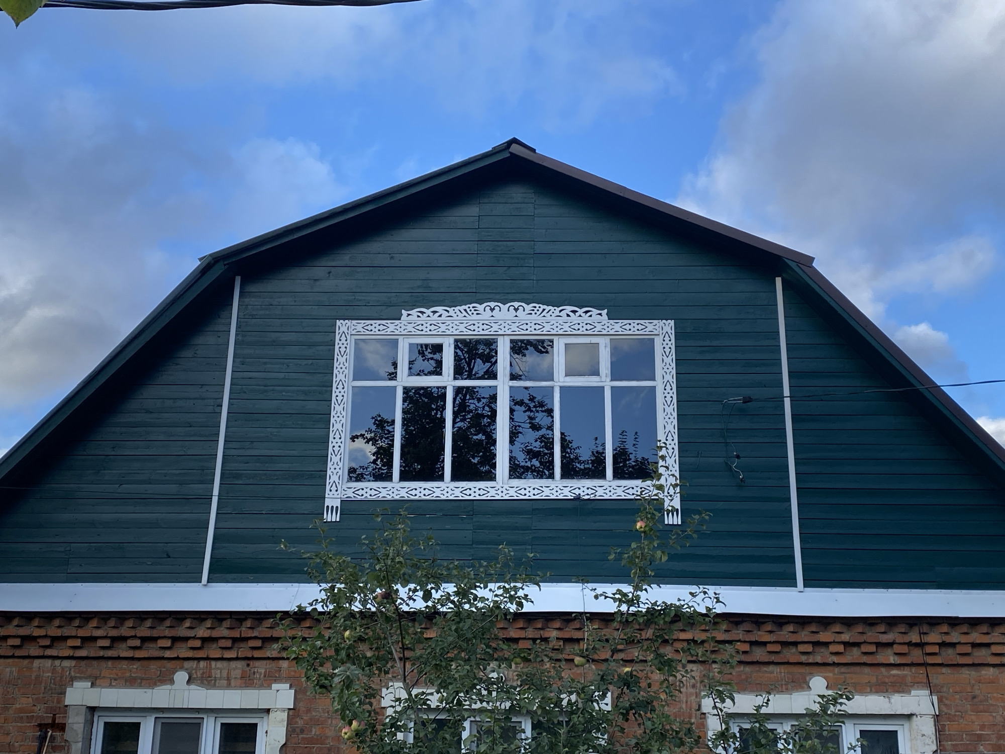 A low-angle, eye-level medium shot captures the upper portion of a rustic, two-story house under a partly cloudy sky. The house features a dark teal wooden clapboard siding on the upper floor, which is gabled. A large, multi-paned window with white trim is centered on the teal section. The window is adorned with intricate white wooden latticework above and on the sides. The lower portion of the house is constructed of red brick. A white trim piece runs horizontally across the top of the brick wall, just below the teal siding. A small tree with green leaves and a few visible red fruits is in the foreground, partially obscuring the lower windows. The sky is a vibrant blue with scattered, fluffy white clouds. A dark wire is visible in the upper left corner of the image.