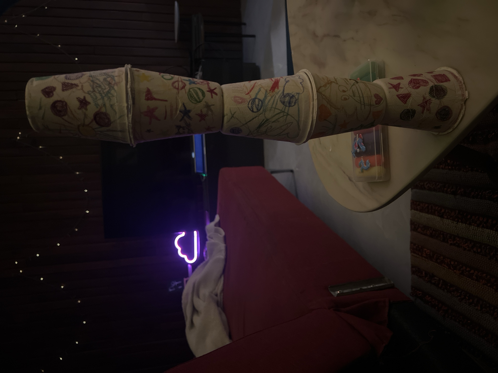 A dimly lit room contains a stack of four paper cups, each adorned with child-like drawings. The cups are stacked vertically on a light-colored, round table. In the background, a dark red couch is partially visible, covered with a white blanket. A purple neon cloud light is mounted on the wall behind the couch. A string of small, twinkling lights is draped across the upper portion of the wall. The scene appears to be indoors, possibly a living room or playroom.