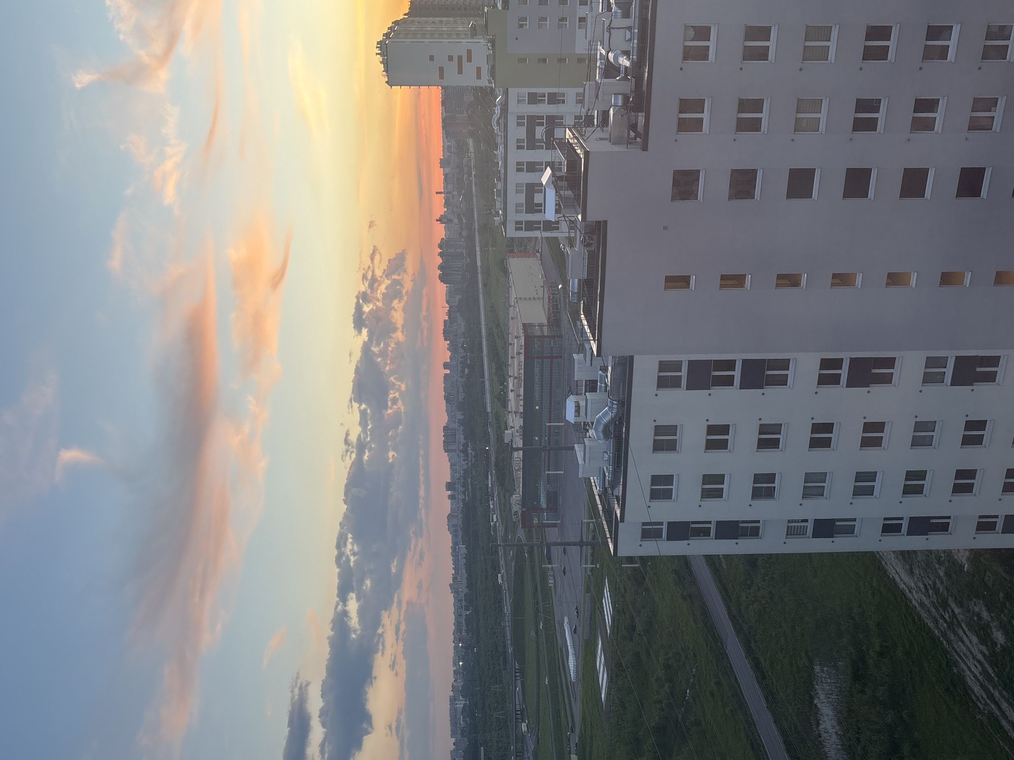 This media file captures an expansive urban or suburban landscape at either sunrise or sunset, characterized by a dramatic sky over buildings and green spaces. The image is oriented vertically but appears to be a landscape shot rotated 90 degrees clockwise.
The upper portion of the scene is dominated by a vast sky showcasing a vibrant color gradient. On the left, it transitions from a soft light blue to shades of peach and pink, deepening into vivid orange and yellow towards the right, indicative of the low sun's position. Scattered clouds, ranging from wispy white to darker grey, are beautifully illuminated with orange hues by the indirect light.
In the midground and foreground, a collection of buildings and land features unfolds. To the right, a prominent light-colored, multi-story building, possibly an apartment or office block, stands tall with numerous square windows arranged in a grid. Some windows reflect light, while others appear dark. Rooftop installations, such as ventilation units or piping, are discernible. Further left and in the distance, other buildings of varying heights and designs, some with green or light grey facades, contribute to the urban backdrop. A faint city skyline is visible on the distant horizon, blending with the colorful sky.
Below the structures, a wide expanse of green grass covers the ground, intersected by a narrow road or path. In the middle distance, a network of roads and indistinct structures suggests an industrial or developing area. No people or specific activities are visible, presenting a static, tranquil scene. There is no clearly readable text within the image. The setting conveys a sense of serene urban sprawl under a magnificent sky, located in Sinyavino, Russia.