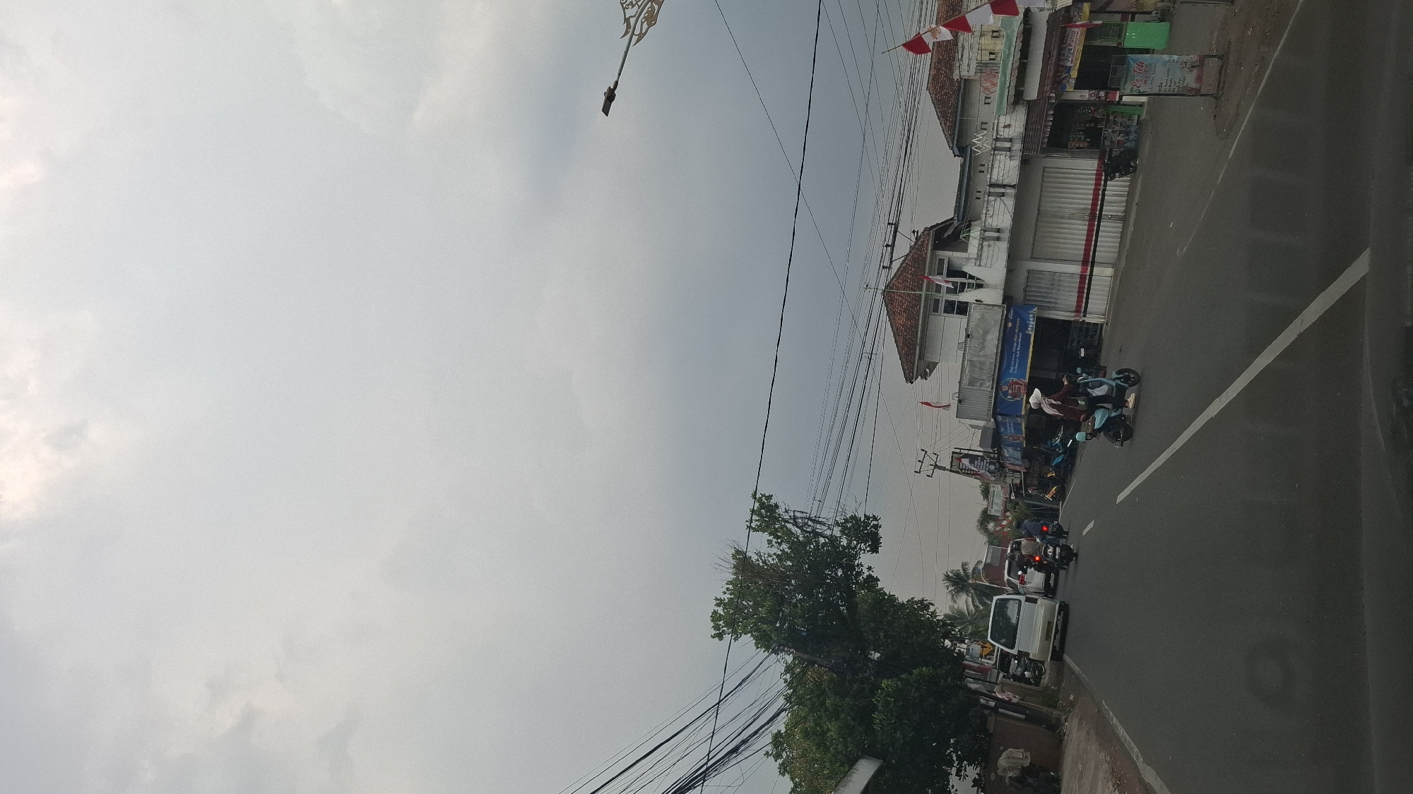 The image shows a street scene in Sukasari, Indonesia, under an overcast sky. The road is paved and has a white lane marker. On the right side of the road, there are several buildings with red-tiled roofs and white walls. One building appears to be a shop with closed shutters. Indonesian flags are visible on some of the buildings. There are many electrical wires strung across the scene.
Several vehicles are present on the road. A white van is parked on the left side of the road. In the center of the road, there are several motorcycles. A person wearing a maroon headscarf and a white hat is riding a light blue electric scooter, traveling away from the viewer. On the left side of the road, there is a large tree with green foliage and some dead branches. In the distance, palm trees are visible. The scene depicts a typical street with commercial establishments and traffic in a residential or semi-urban area.