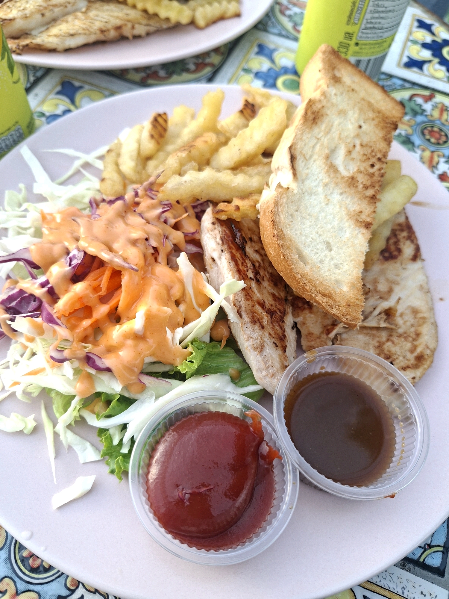 The image shows a close-up of a meal served on a pink plate, with a tiled surface and a portion of a yellow bottle visible in the background. The meal consists of grilled chicken breast, french fries, a salad with shredded carrots and red cabbage topped with a creamy orange dressing, and two small containers of dipping sauces – one with ketchup and the other with a dark brown sauce. The grilled chicken appears to have grill marks. A slice of toasted bread is also placed on top of the chicken. The lighting suggests an outdoor or brightly lit indoor setting. The overall scene depicts a casual dining experience. While the exact location in Pattaya City, Thailand is not specified, the presence of a colorful tiled surface and the style of the food and drinks are consistent with casual eateries found in tourist areas.