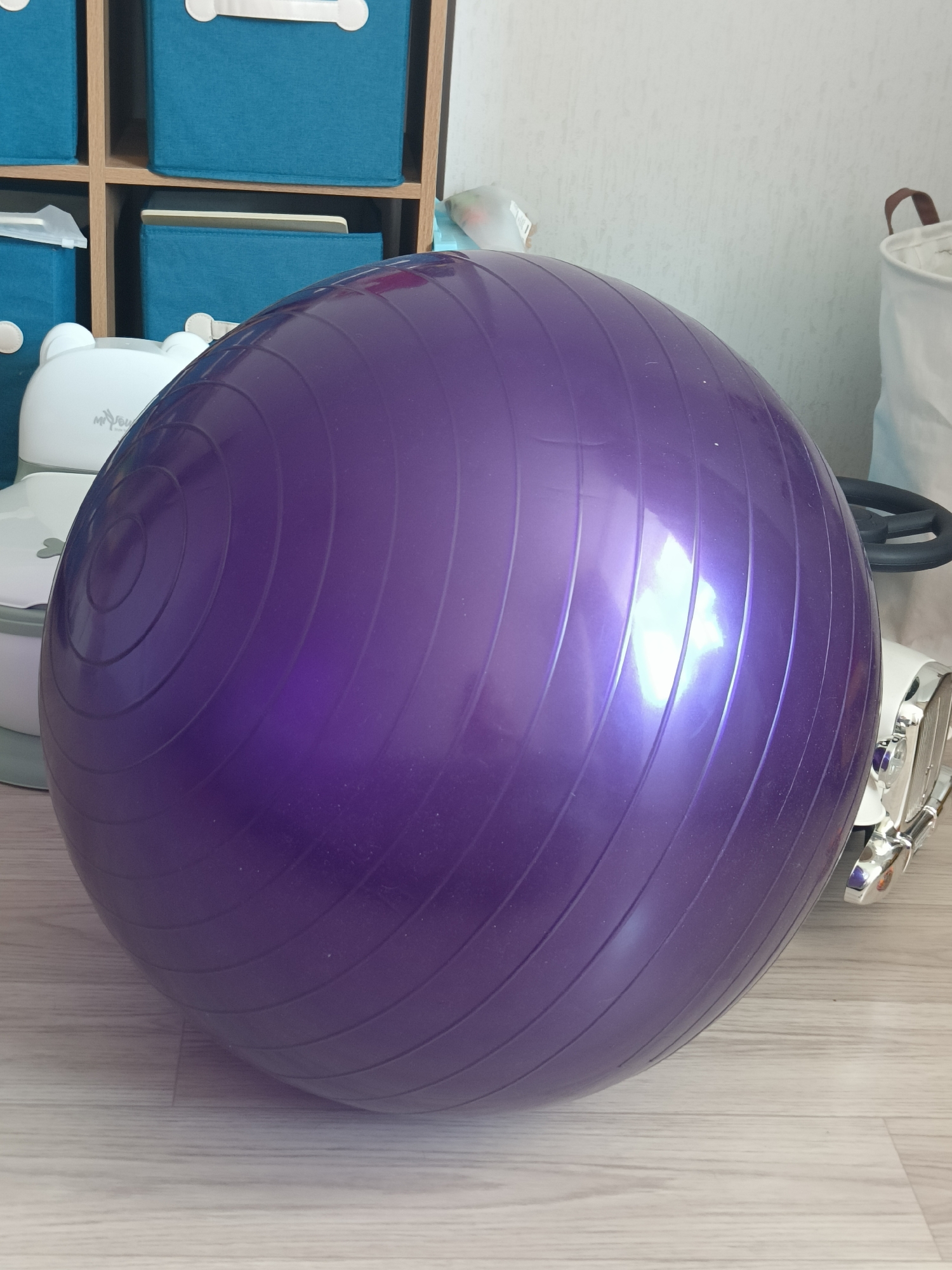 A large, purple exercise ball is the central focus of the image, positioned on a light-colored wooden floor. The ball has a smooth, shiny surface with visible concentric lines molded into it, indicating its texture and design. Reflections of light are prominent on its surface, suggesting an indoor setting with artificial lighting.
In the background, to the left of the exercise ball, is a shelving unit with several blue fabric storage cubes. Some of these cubes have white handles. Partially visible behind the exercise ball are white objects that appear to be baby-related items, one with a bear-like design on top and another that resembles a potty or a small chair with a lid. Further to the right, a white fabric basket with a brown handle is visible against a textured white wall. There are also glimpses of a white object with chrome accents and a black steering wheel, which could be a toy car or a similar item.
The scene appears to be an indoor domestic space, possibly a playroom or a nursery, given the presence of the baby items and storage solutions. The lighting is even, and there are no strong shadows, suggesting it is not a specific time of day that is clearly indicated by natural light. No people are visible in the image. The overall impression is one of a tidy, organized living area. No visible text is discernable in the image.