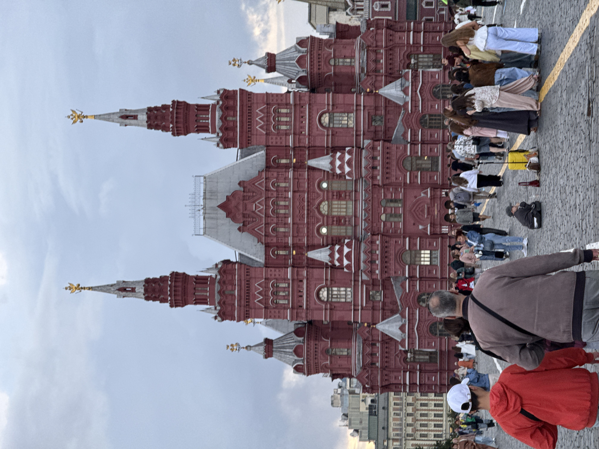 This media file captures a segment of a grand, red-brick building with ornate architectural details, possibly a historical landmark. The building features multiple towers with pointed roofs, some adorned with golden spires topped with winged figures. The facade is characterized by numerous windows, decorative brickwork, and varied rooflines.
In the foreground, a group of people are visible, appearing to be tourists or visitors. They are standing on a cobblestone ground, with a prominent yellow line marking a path or boundary. The people are casually dressed, suggesting a daytime activity. Some are looking towards the building, while others are interacting amongst themselves. One person in a brown jacket is facing away from the camera, while another in a red jacket with a white baseball cap is also oriented away.
The scene is set outdoors, under a slightly overcast sky with visible clouds. The lighting suggests it is likely daytime, possibly late afternoon or early evening given the softer light. The red architecture and cobblestone ground are strong cues for a location like Moscow, Russia, as indicated. There are no visible text elements within the image. The overall atmosphere appears to be one of exploration and appreciation of the historical architecture.