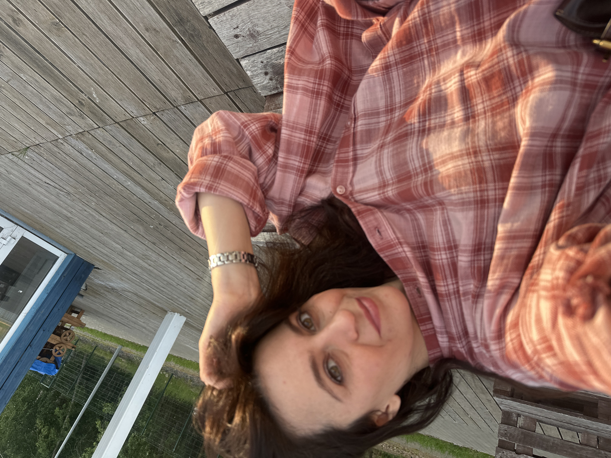 The image displays a woman taking a selfie outdoors in Chaikovsky, Russia, characterized by warm, low-angle sunlight consistent with late afternoon or early evening. The woman has dark hair and brown eyes, looking directly at the camera with a subtle smile. She wears a pink and white plaid flannel shirt, with her left hand touching her hair. A silver-colored link bracelet or watch is visible on her left wrist. She is seated or leaning on a weathered wooden deck composed of horizontal planks. In the background, to the viewer's left, a wooden pallet-like structure is visible. Towards the right, a tall white pole stands, and a blue-painted building with a white-framed window or door is present. Further back, beyond a wire fence, green foliage and trees are visible, along with a decorative wooden cart, partially covered by a blue tarp. The ground behind the deck transitions to a paved area before reaching the fence line.