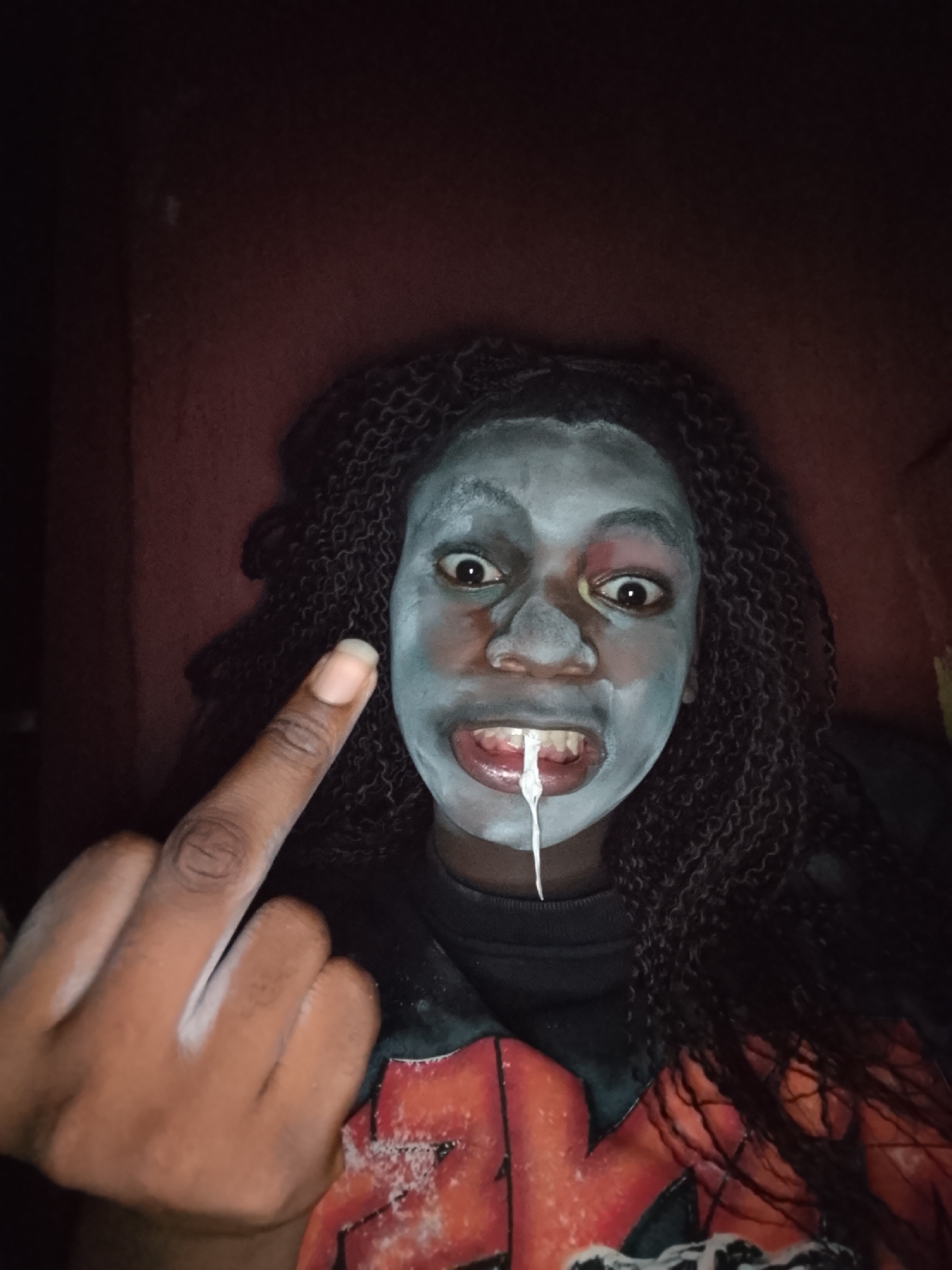 A dimly lit, close-up portrait photograph captures an individual in an indoor setting, likely in Onitsha, Nigeria. The person, with dark skin and long, dark, braided or curly hair, looks directly at the camera with wide, intense eyes. Their face is partially covered in a light blue or grey substance resembling face paint or a mask, with reddish smudges around the eyes, suggesting a distressed or artistic makeup effect.
From the person's open mouth, a thick, white, stringy substance drips downwards, creating a drool-like or slime-like appearance that contributes to a grotesque or horror-themed aesthetic. The individual wears a dark garment, possibly a t-shirt or hoodie, with a distinct reddish-orange graphic design visible on the chest. Their left hand is raised, with the middle finger prominently extended towards the viewer in a defiant or rude gesture, and traces of the white substance are also visible on their fingers.
The background consists of a dark red or maroon wall, obscured by shadows, indicating low light conditions. The overall mood of the image is unsettling, defiant, and theatrical, suggesting a posed shot for artistic expression, costume, or a playful horror theme. No specific location cues are visible within the image to confirm Onitsha, Nigeria, beyond the provided context. There is no visible text.