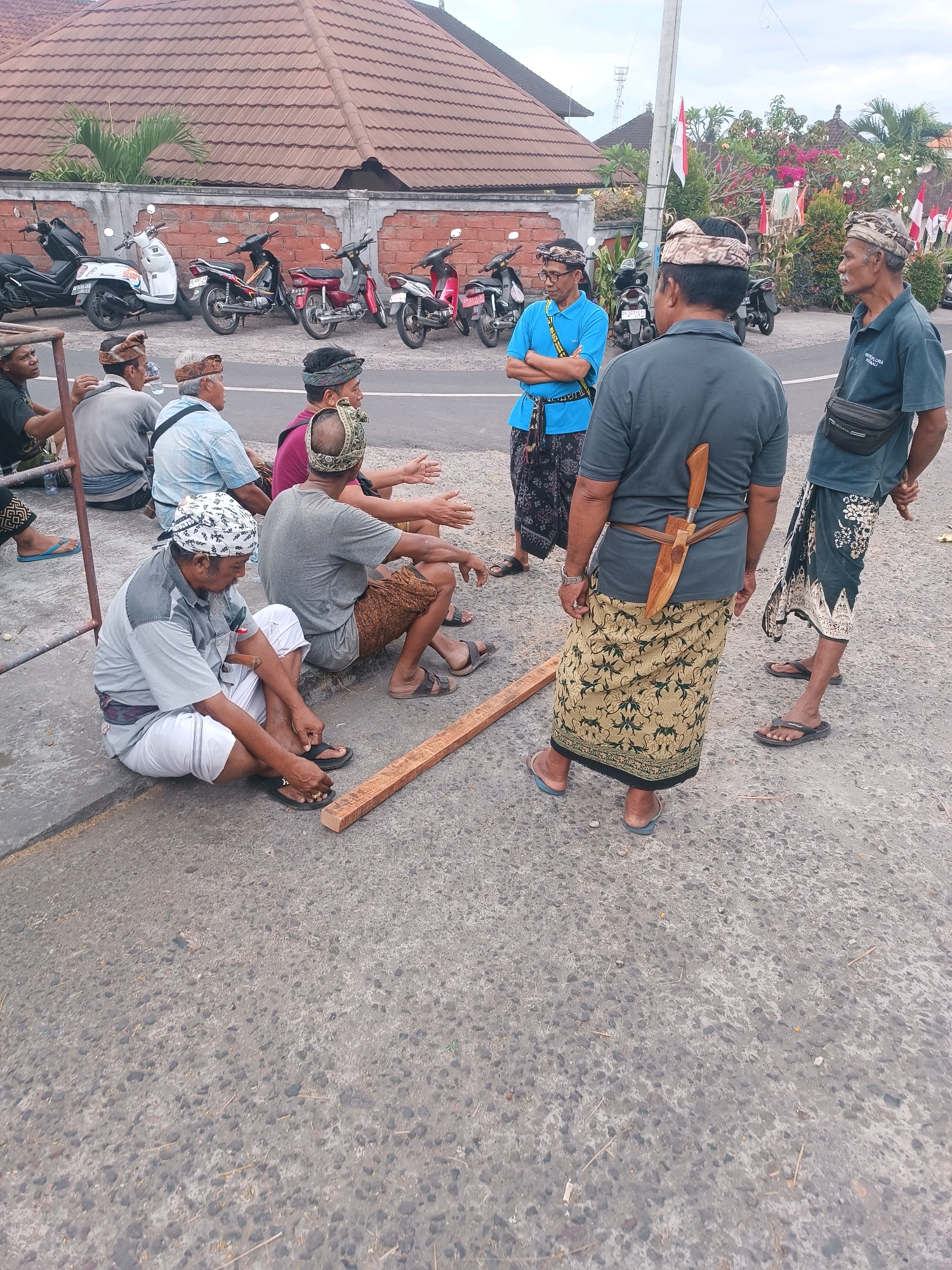 In Selemadeg, Indonesia, a group of men are gathered on a paved road, some seated and others standing. The men are wearing traditional Balinese attire, including headbands (udeng) and sarongs. One man, standing and facing away from the camera, has a large, ornate dagger (keris) tucked into his sarong at his back. A piece of lumber lies on the ground in front of some of the seated men. Behind them, motorcycles are parked next to a building with a tiled roof. The environment appears to be a roadside setting, with a curb visible along the edge of the pavement. The sky is overcast, and flags are visible in the background, suggesting a festive or communal occasion.