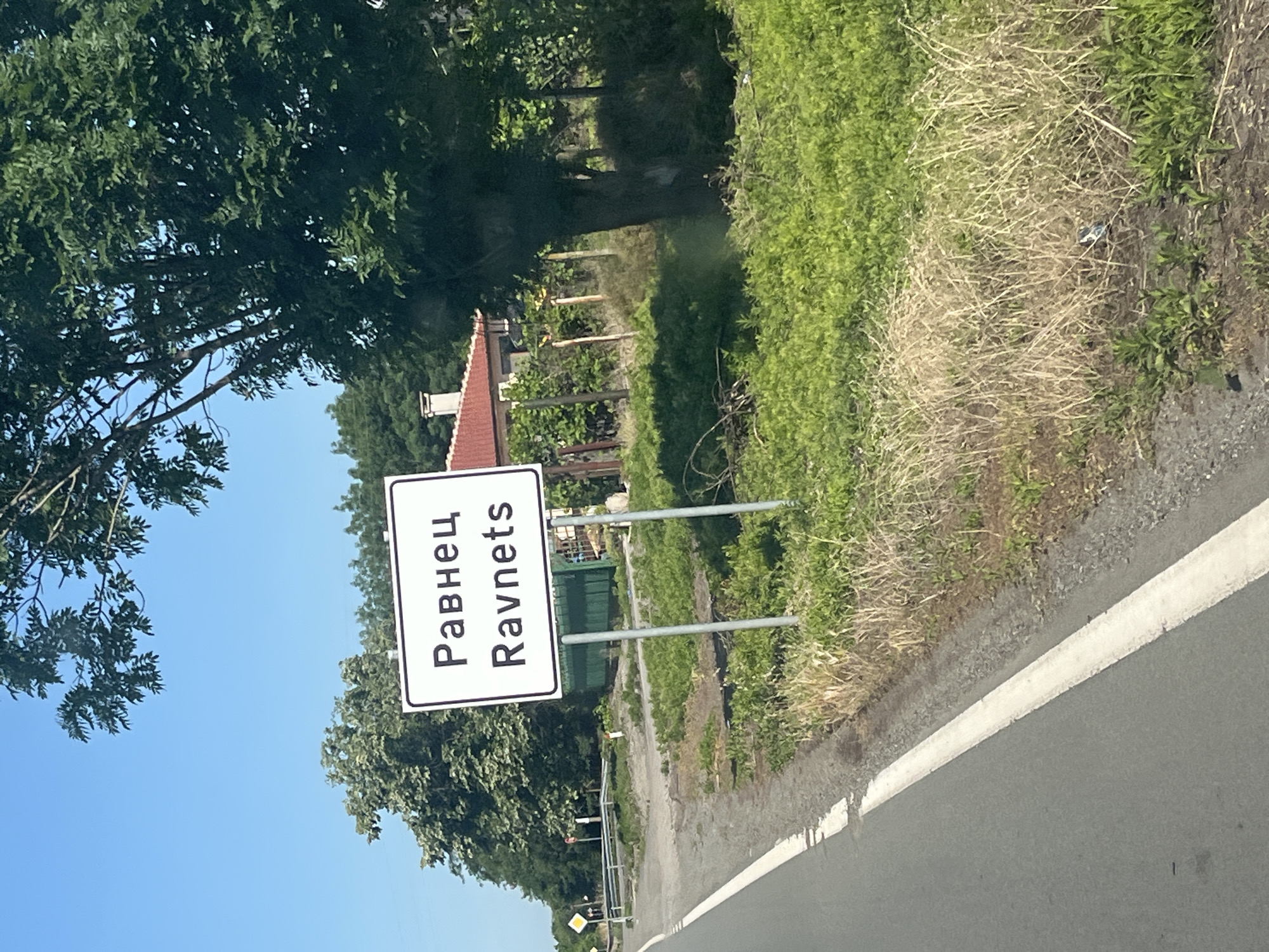 The image shows a street sign indicating the location of Ravnets, Bulgaria. The sign, which is white with black lettering, reads "Равнец" in Cyrillic and "Ravnets" in Latin script. It is mounted on two metal poles. The sign is situated on the side of a paved road, with a grassy area and trees in the background. A red-roofed building is partially visible behind the trees. The sky is blue and clear. The overall scene depicts a rural or suburban entrance to the town.