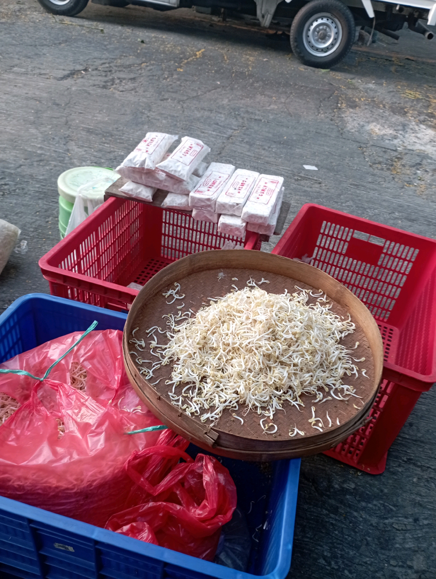 The image depicts an outdoor market scene in Ponorogo, Indonesia. In the foreground, a blue plastic crate contains several red plastic bags filled with white bean sprouts. To the right of the crate, a woven wooden tray is piled high with more bean sprouts. Behind this, two red plastic baskets are visible, one partially filled with white packaged goods that appear to be tempeh, each wrapped in paper with red text. In the background, a white utility vehicle is parked on a paved surface, and a green and white plastic container is also present. The ground is a rough, gray asphalt or concrete. The overall impression is of a food stall preparing for or in the midst of selling produce.