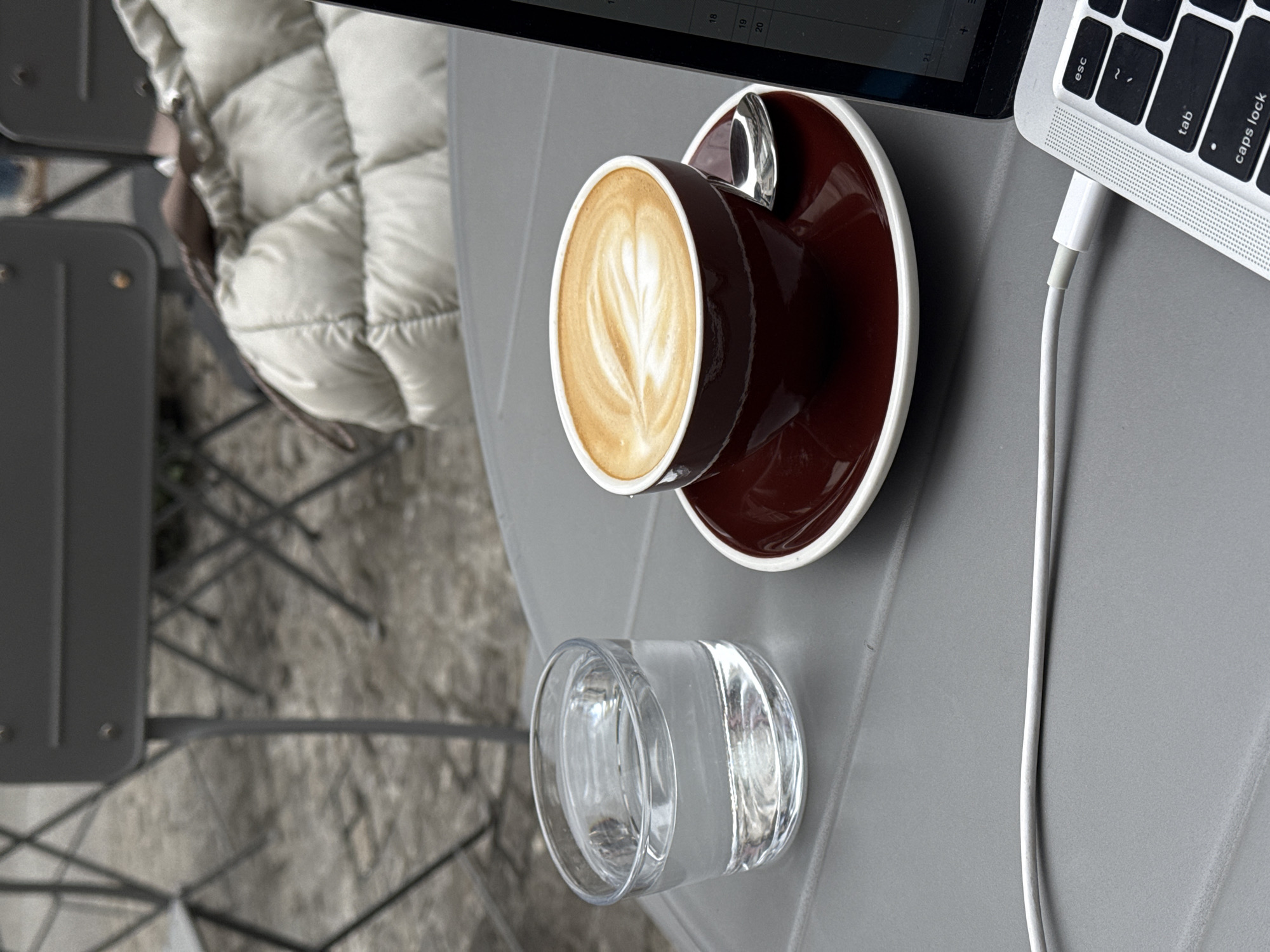 A close-up shot of a grey table in an outdoor setting, likely in Belgrade, Serbia, given the context. On the table is a laptop, a brown ceramic coffee cup with latte art on a saucer, a glass of water, and a white charging cable connected to the laptop. Part of a quilted jacket is visible in the background, suggesting someone is seated nearby. A metal chair is also partially visible. The scene suggests a casual outdoor cafe or workspace environment.