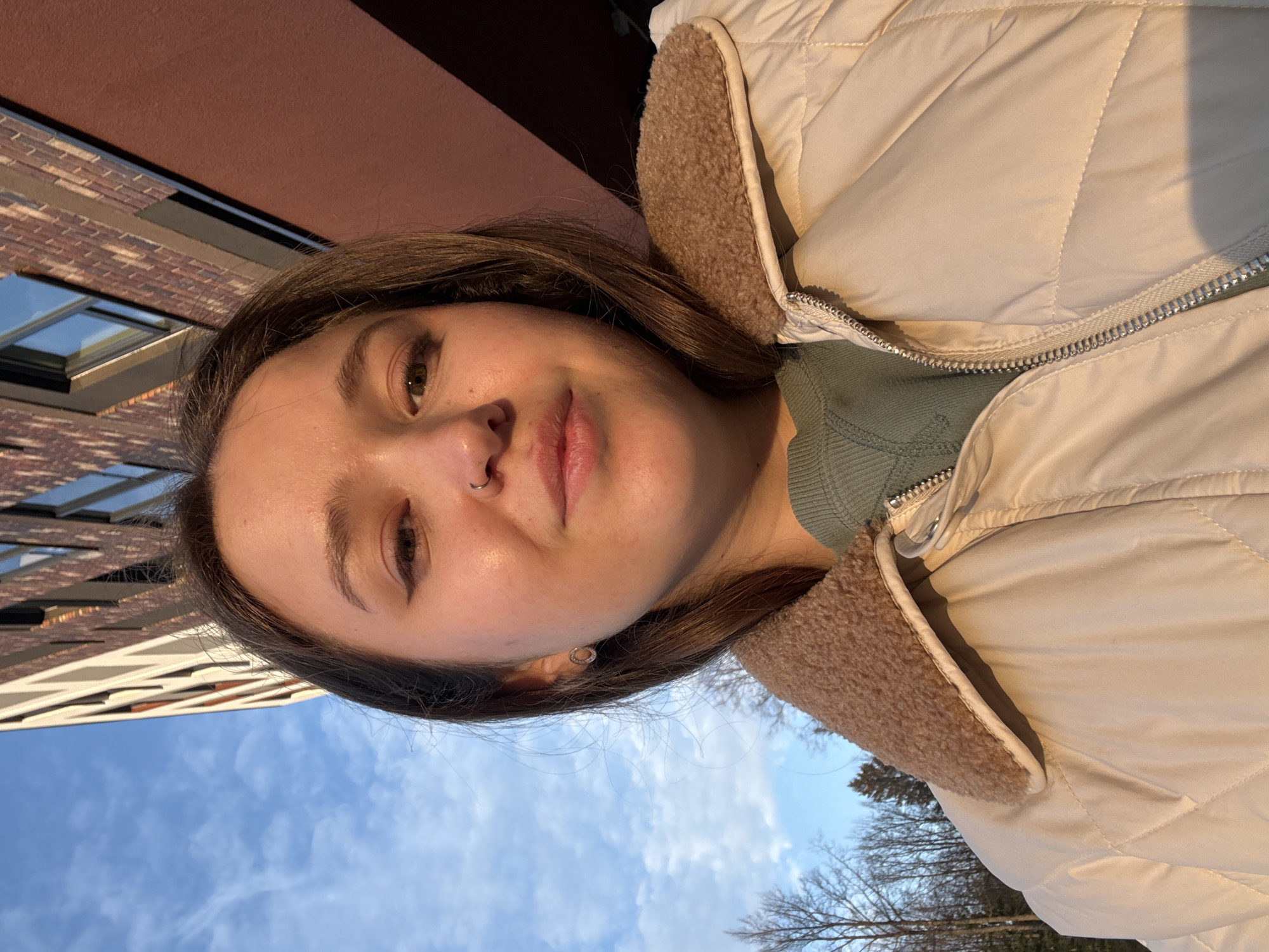 A young woman with brown hair and a nose piercing is centered in the frame, facing the camera. She wears a beige puffer jacket with a fuzzy collar and a green top underneath. Behind her, the sky is visible with thin, white clouds. To the left, a building with a brick facade and visible windows rises into the sky. To the right, bare tree branches suggest either late autumn or early spring. Based on the architectural style, the location could potentially be Izhevsk, Russia, but further information would be needed to confirm.