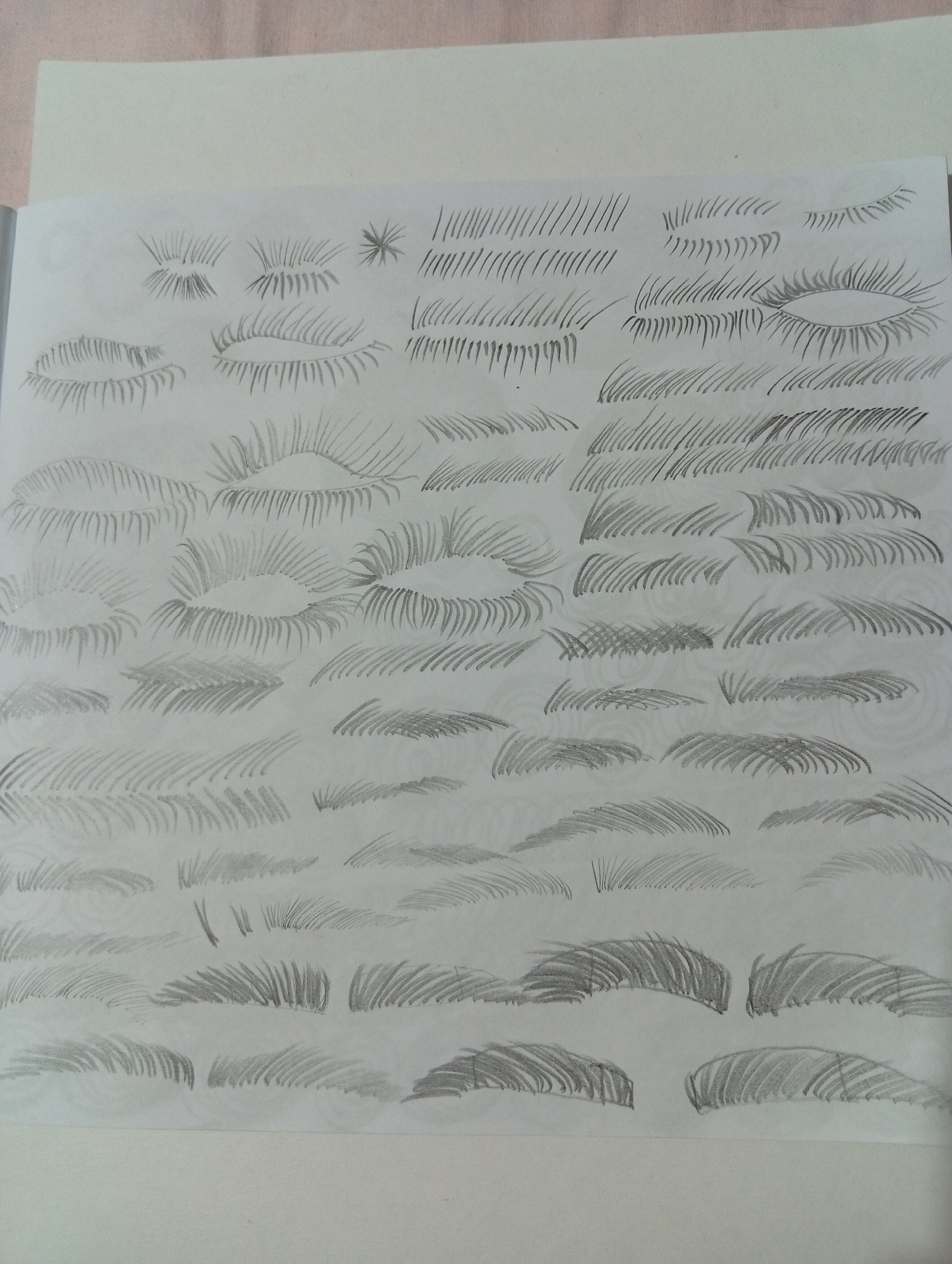 The image shows a sketchbook page filled with pencil drawings of eyelashes and eyebrows. The drawings are varied in style and density, demonstrating different techniques for rendering these features. Some sketches depict individual eyelashes, while others show full sets of lashes and eyebrows. The page is laid flat, with the top edge of the sketchbook cover visible. The background is a plain surface, possibly a table or desk. The image is a close-up of the sketchbook, with no discernible background elements to indicate a specific location, including San Antonio de Los Altos, Venezuela.