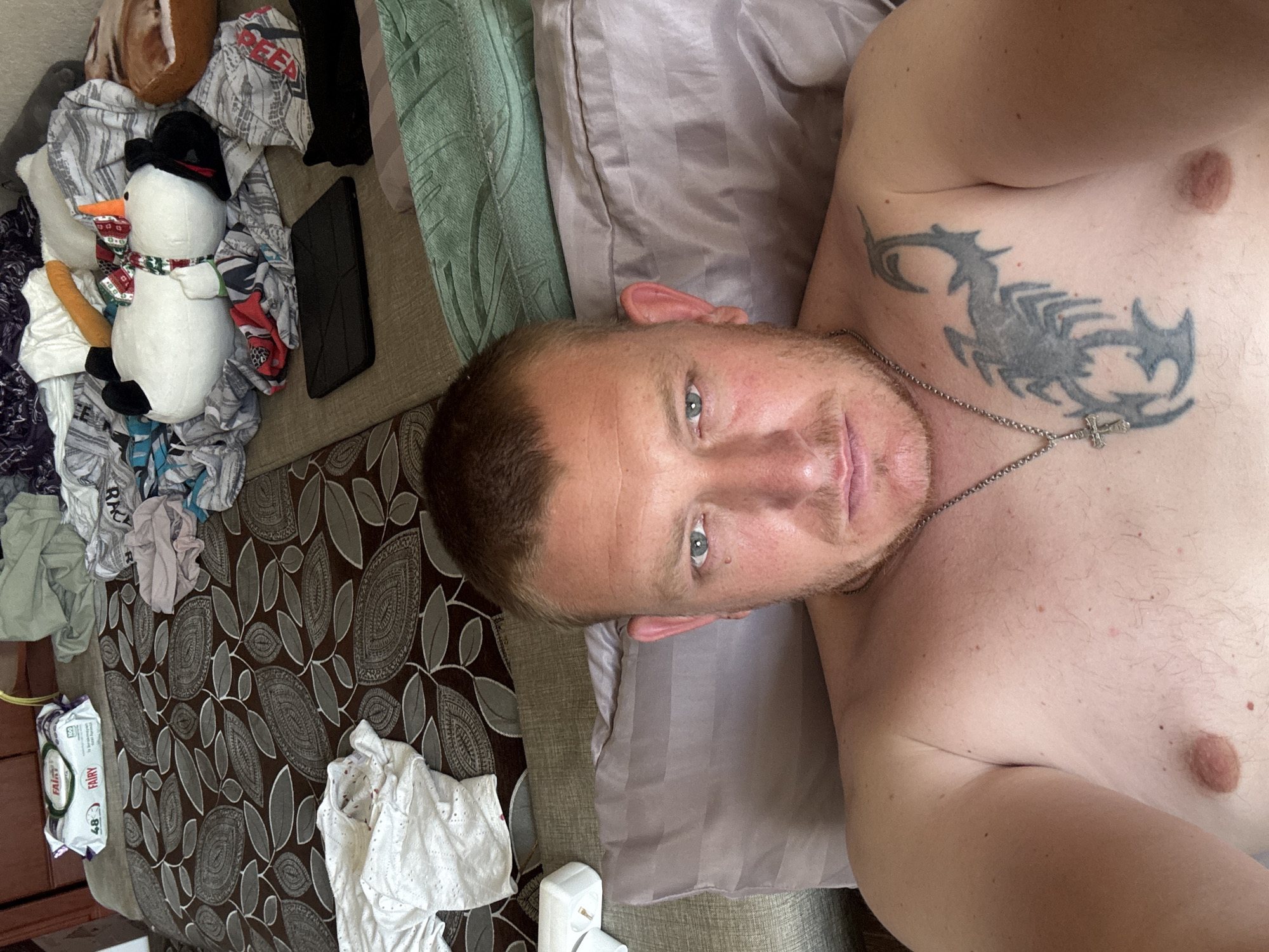 An individual lies on a bed, their upper body visible. The background is cluttered with various items, including a patterned bedspread, scattered clothing, a plush snowman toy, and what appears to be baby wipes. The individual has a scorpion tattoo on their chest and is wearing a silver chain with a cross pendant. The lighting suggests an indoor setting, possibly during the day.