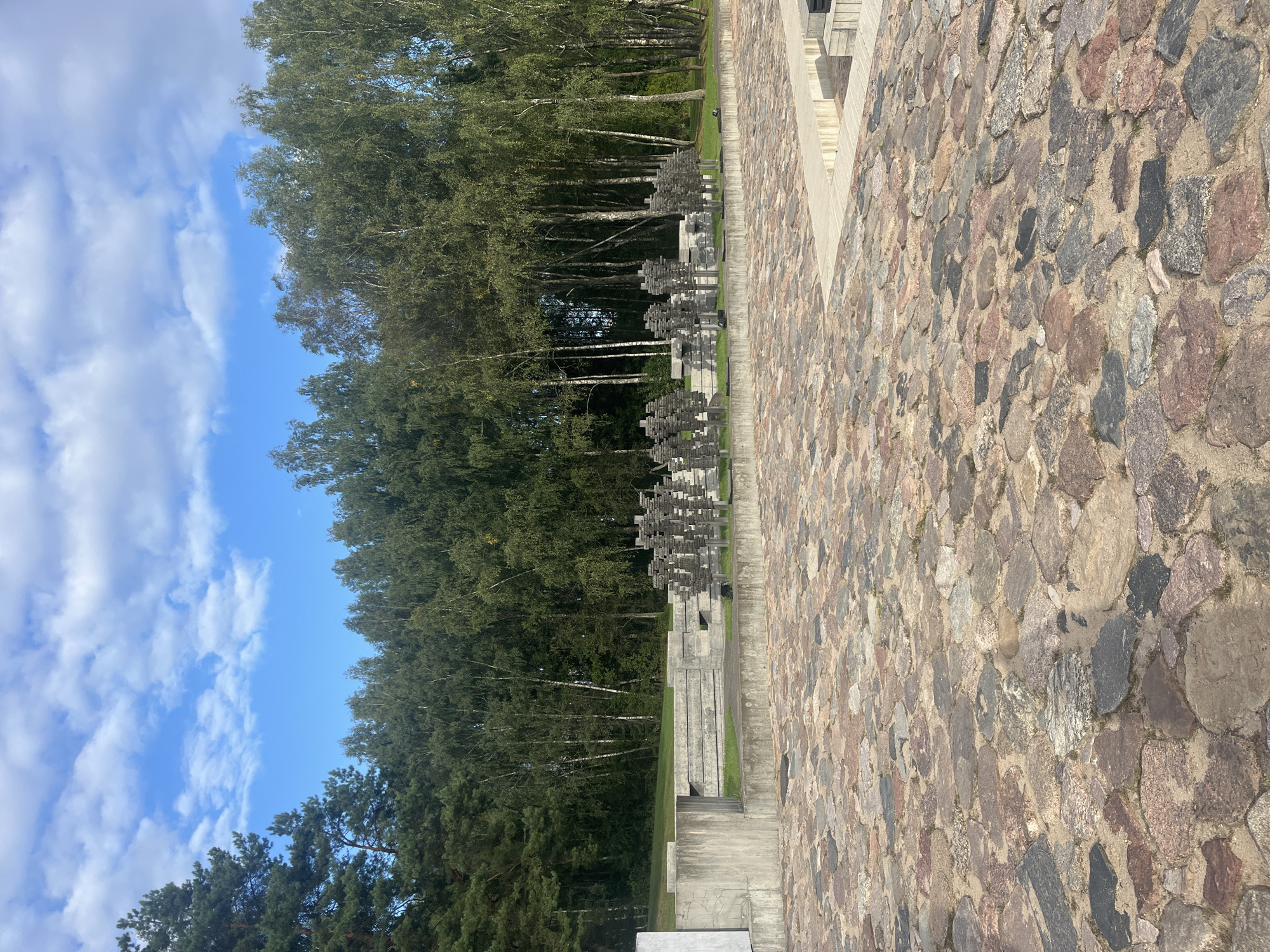 The image shows an outdoor memorial site. In the foreground is a paved area made of large, irregular-shaped stones in various shades of pink, gray, and brown. Behind this area, a concrete structure with tiered levels and horizontal railings is visible. To the right of the concrete structure, a series of dark, geometric sculptures resembling abstract figures stand in rows. These sculptures are mounted on poles and are arranged in front of a dense line of trees, primarily birch and pine. The trees form a natural backdrop against a bright blue sky with scattered white clouds. The overall impression is one of a solemn, open-air monument.
