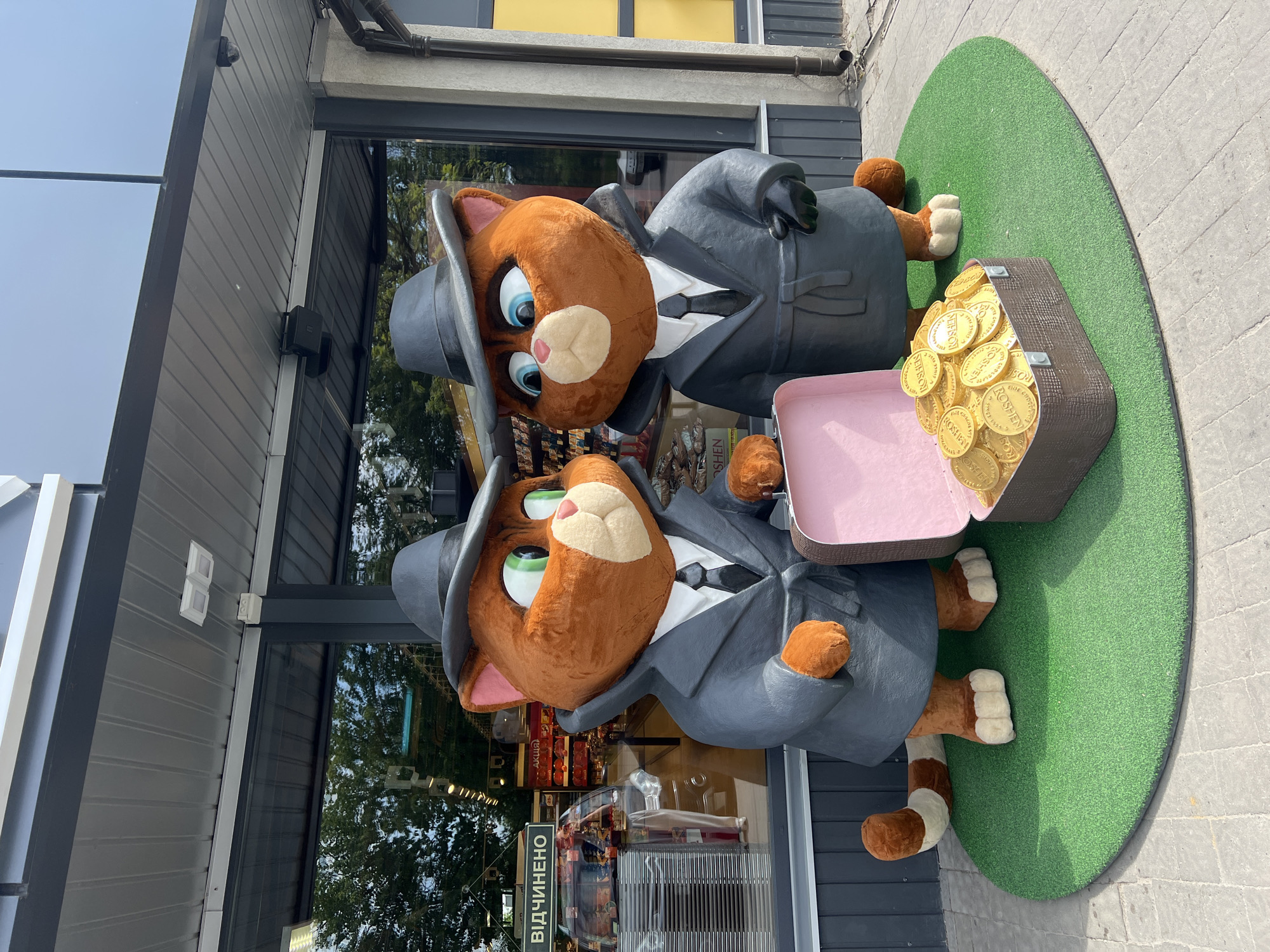 Two cartoon cat figures dressed as gangsters stand on a green circular mat in front of a building. Each cat wears a gray suit with a white shirt and a black tie, as well as a gray fedora. One cat has blue eyes, the other has green. They are positioned next to an open suitcase filled with gold-colored coins. The coins bear the "ROSHEN" logo. The building behind the figures has a reflective glass facade, showing the interior of what appears to be a store, potentially a confectionery given the Roshen branding. A sign in Ukrainian, "ВІДЧИНЕНО" (meaning "Open"), is visible on the door. The location is likely a Roshen store in Ternopil, Ukraine, based on the presence of the figures and products. The exterior flooring is concrete, and the building has gray siding and a yellow accent above the store window.