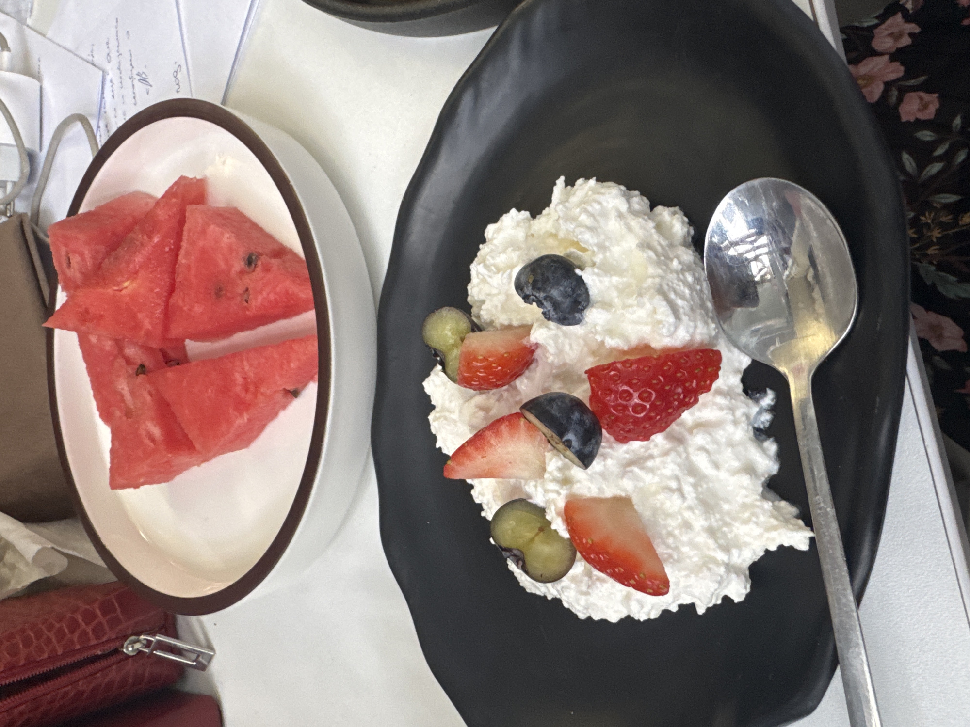 The image shows a close-up view of a dessert and a side dish of watermelon. On the left, a white bowl contains several triangular slices of watermelon. On the right, a black plate holds a dollop of whipped cream topped with various fruits, including strawberries, blueberries, and grapes. A silver spoon rests on the plate, partially submerged in the whipped cream. The background appears to be a table with some papers and a textured fabric, possibly clothing with a floral pattern. The lighting suggests it is daytime. There is no discernible text in the image.