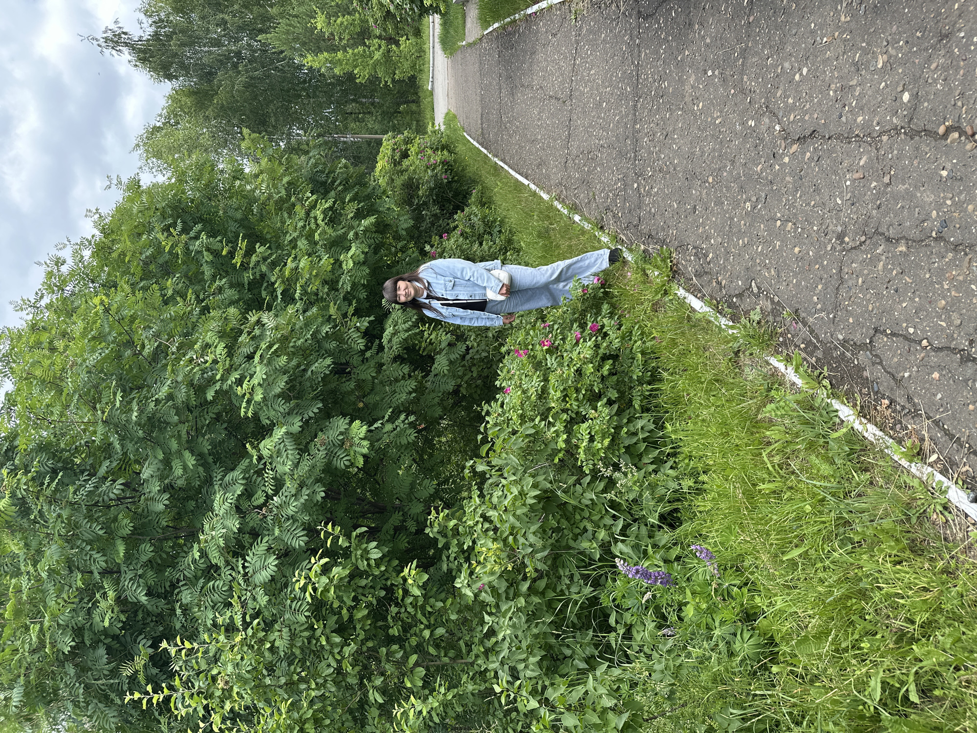 A young woman stands on the right side of a paved pathway, surrounded by lush green foliage. She is wearing a light blue denim jacket, a black top, and light blue jeans. A white purse is slung across her shoulder. The pathway appears to be in a park or wooded area, with tall trees and bushes lining its edge. The ground is covered in grass and wildflowers, including purple lupine and pink roses. The sky is overcast with clouds. The location is the park near the Kama River in Chaikovsky, Russia.