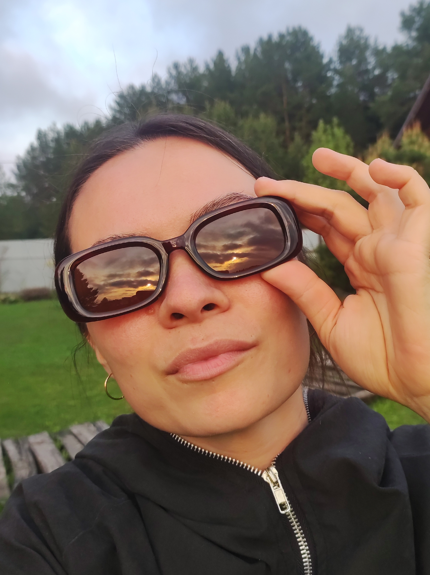 A woman is holding her sunglasses up to her face outdoors. The reflection on the sunglasses shows a sunset. The woman has dark hair, a light complexion and is wearing a black hooded jacket. She has a small gold hoop earring in her left ear. There are trees in the background. There is no information to identify the location in Фоки, Russia.