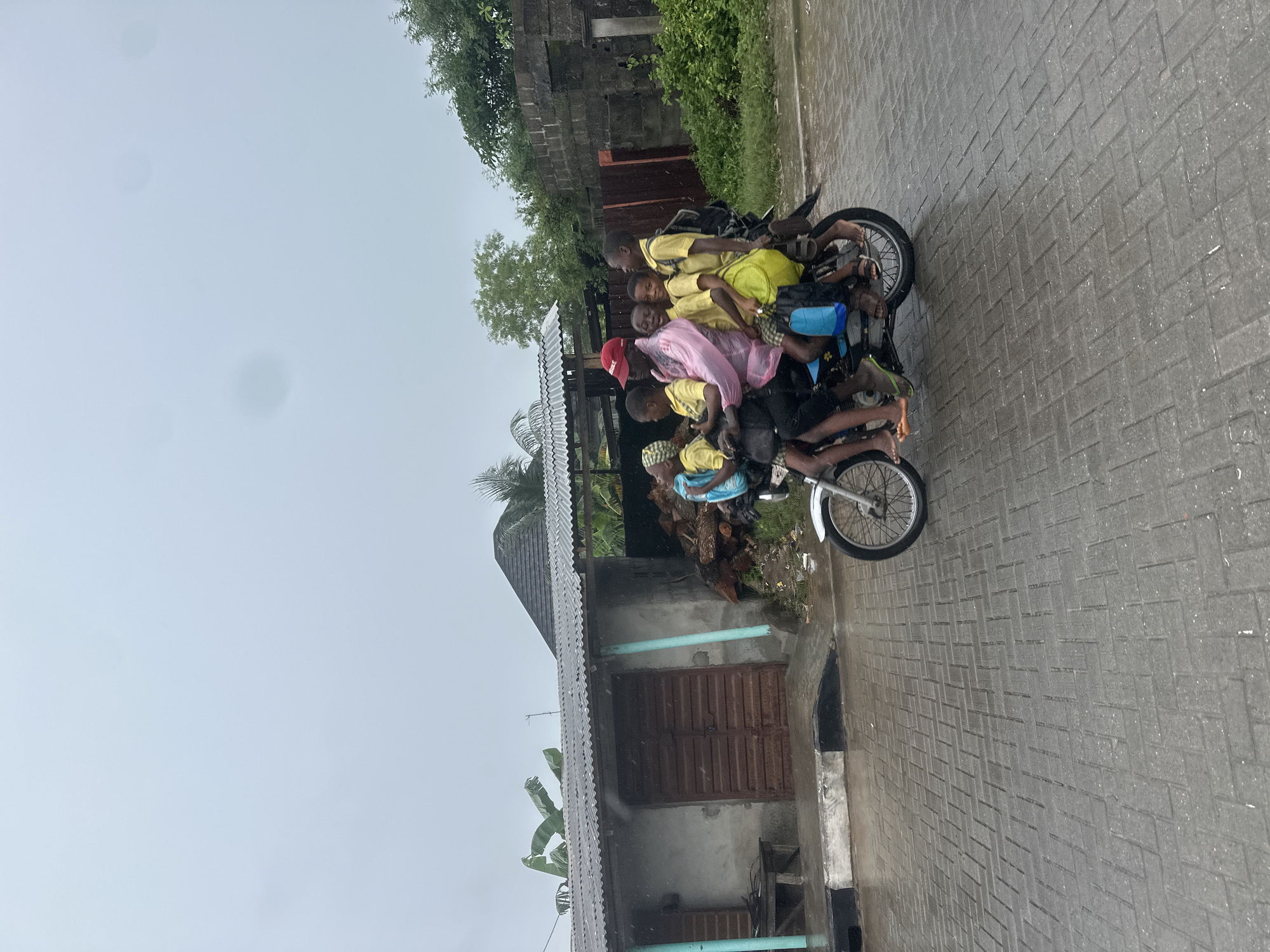 The image shows a group of approximately nine children, likely students, crammed onto a single motorcycle. They are all wearing yellow shirts, suggesting they are in uniform. Some children are perched in front of the rider, while others are squeezed behind him. One child is wearing a red cap, and another is partially covered by a pink plastic sheet. The children's faces are visible, and their expressions suggest a mix of composure and perhaps some discomfort due to the crowded situation.
The motorcycle is on a wet, paved sidewalk, indicating it has recently rained or is currently raining. The weather appears overcast and gloomy. The background includes a building with a corrugated metal roof, a wooden door, and some greenery. There are also hints of more substantial structures with stone elements and vegetation in the distance. The setting appears to be a residential or semi-urban area in Badagry, Nigeria. The wet pavement and the way the children are dressed suggest a humid climate. The overall scene depicts a common, albeit potentially unsafe, mode of transportation for a large group of children. No visible text is present in the image.