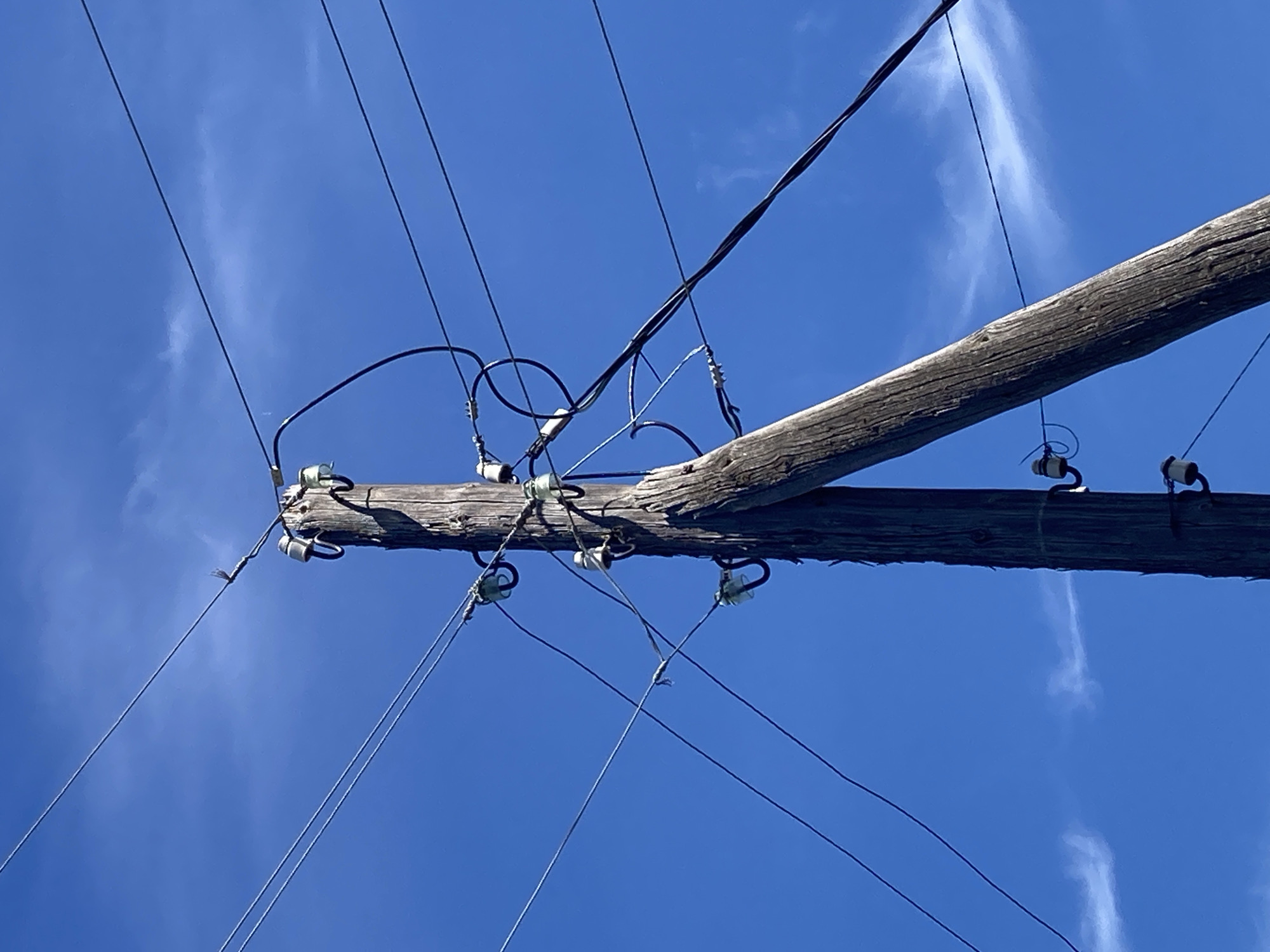 A weathered wooden utility pole, leaning slightly to the right, stands against a bright blue sky with sparse white clouds. The pole is adorned with multiple ceramic insulators, each holding at least one dark electrical wire. These wires fan out from the pole in various directions, creating a complex network against the sky. Some wires appear to be twisted together. The pole itself shows signs of age, with a rough, textured surface. The overall scene suggests an outdoor setting, likely in a rural or semi-rural area given the rustic nature of the pole and the clear sky.