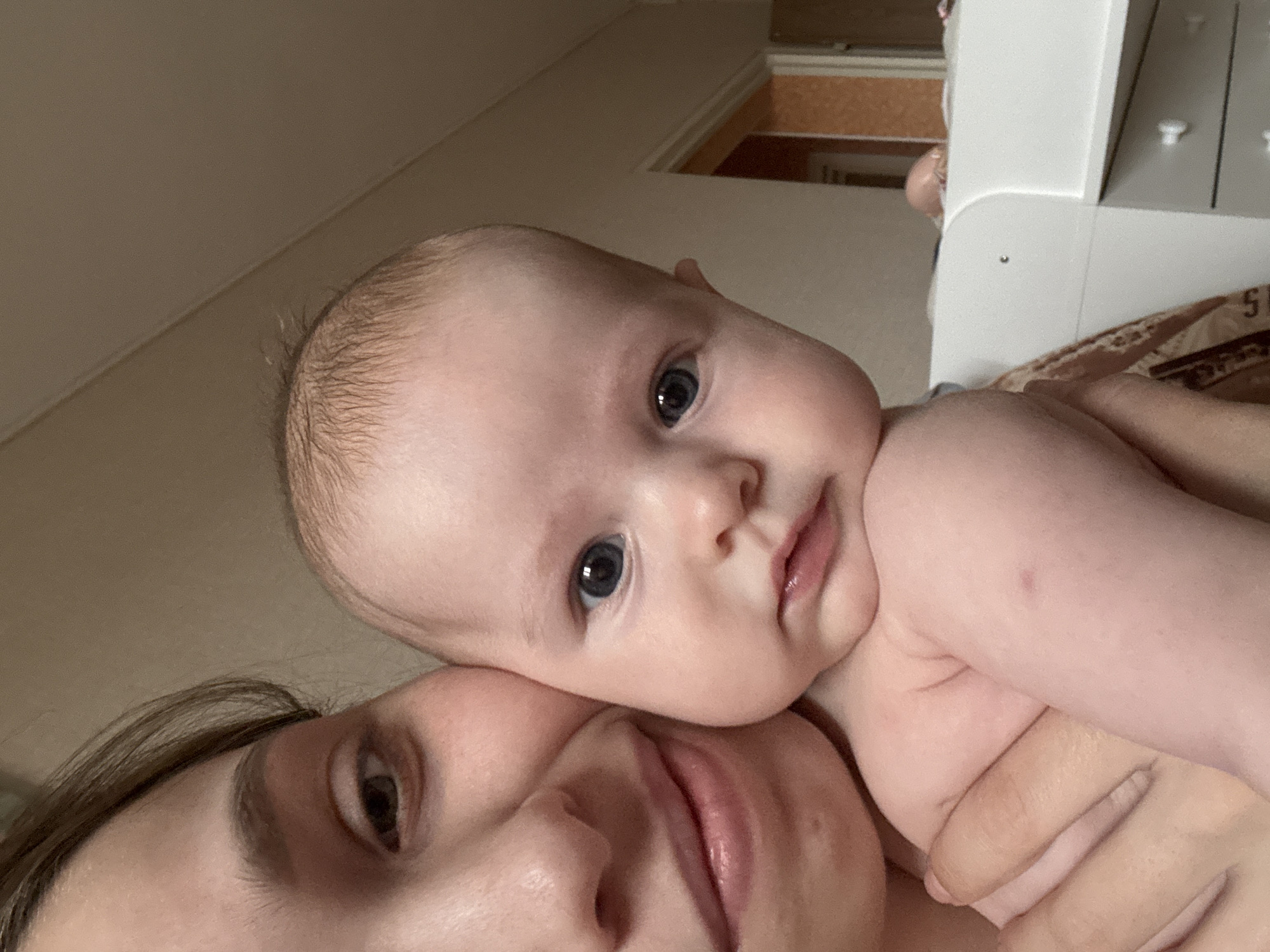 This intimate indoor photograph, taken in Chaikovsky, Russia, captures a close-up, slightly angled view of a mother and her baby, likely a selfie. The mother's face is partially visible in the lower left, showing her forehead, a gentle dark eye looking towards the camera, and part of her lips and chin, framed by dark brown hair.
The baby's head and upper torso dominate the center and upper right of the frame, resting affectionately on the mother's head or shoulder. The baby has fine, light hair and captivating dark blue eyes that gaze directly at the viewer. Its fair skin and plump features are evident, with a small reddish mark visible on its right arm or shoulder. The baby is partially shirtless, revealing its bare shoulder.
The background consists of a plain, light brown or beige wall/ceiling in the upper left. In the upper right, a white piece of furniture, possibly a crib or bed frame, shows structural details like small white knobs and a dark vertical line. Behind this furniture, a patterned fabric, likely a blanket or sheet, displays a partial number "5" and another indistinct character. Further back, a hint of a light brown or orange textured surface is visible.
The soft, even lighting suggests an indoor setting. The image conveys a tender and affectionate moment between mother and child, with the baby's expression appearing curious and direct, and the mother's expression gentle. No other specific location or time-of-day cues are discernible from the visual information.