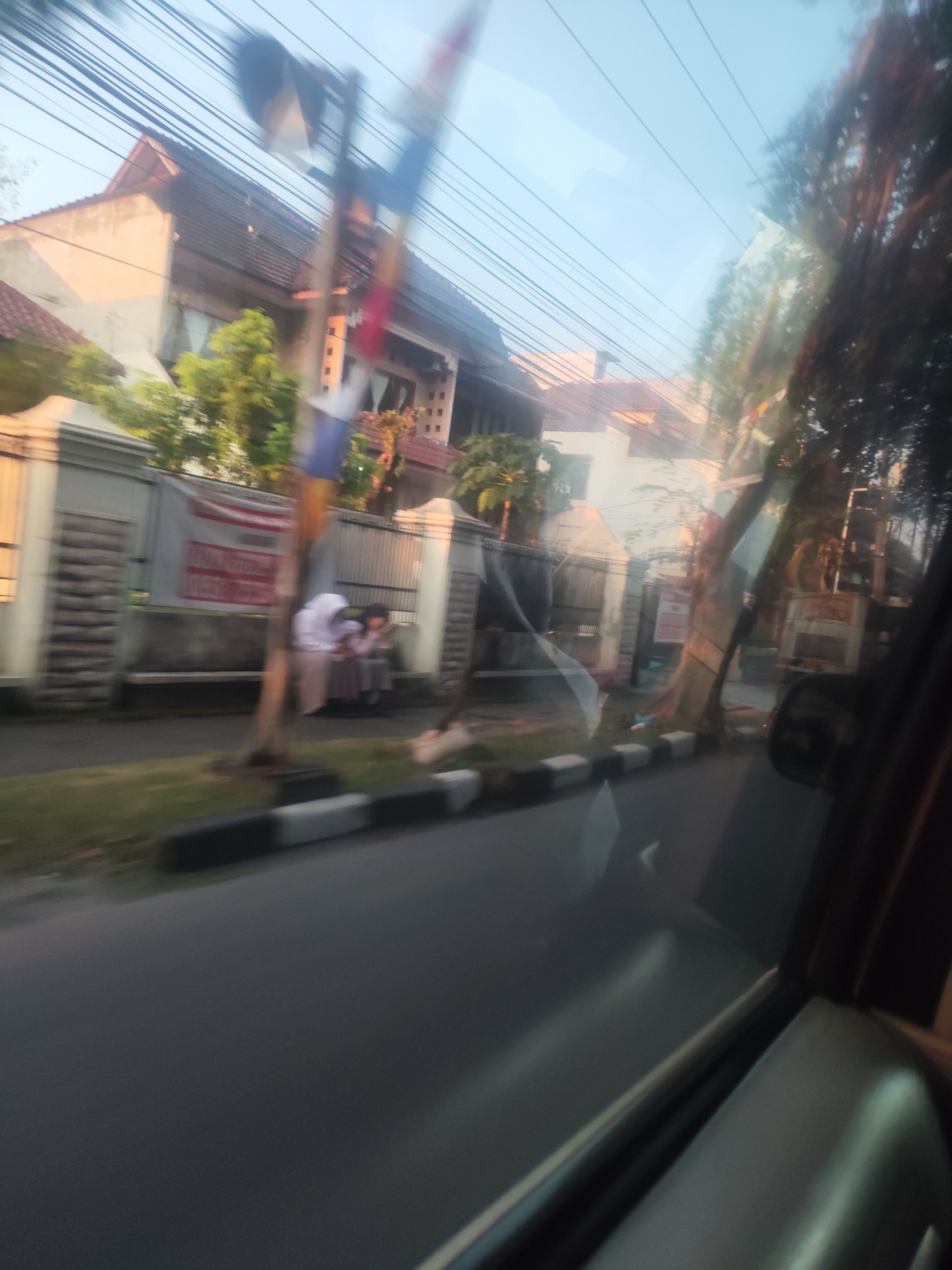 The image captures a street scene in the City of Medan, Indonesia, viewed through a car window. The perspective is from inside a vehicle, indicated by reflections on the glass and the presence of a car interior element in the foreground right. The scene shows a row of houses with beige and orange facades, partially obscured by lush green trees and a large, mature tree with a thick trunk situated near the road. A grey fence with vertical slats runs along the front of the properties. Two individuals, possibly students wearing light-colored uniforms, are seated near the fence. One appears to be wearing a hijab. A banner with red and white lettering is visible behind them. Power lines crisscross the sky. The road in the foreground is paved, with a black and white striped curb separating it from a grassy area. The overall impression is of a residential street during daylight hours, with a slightly hazy or blurred quality, suggesting the image was taken while in motion.