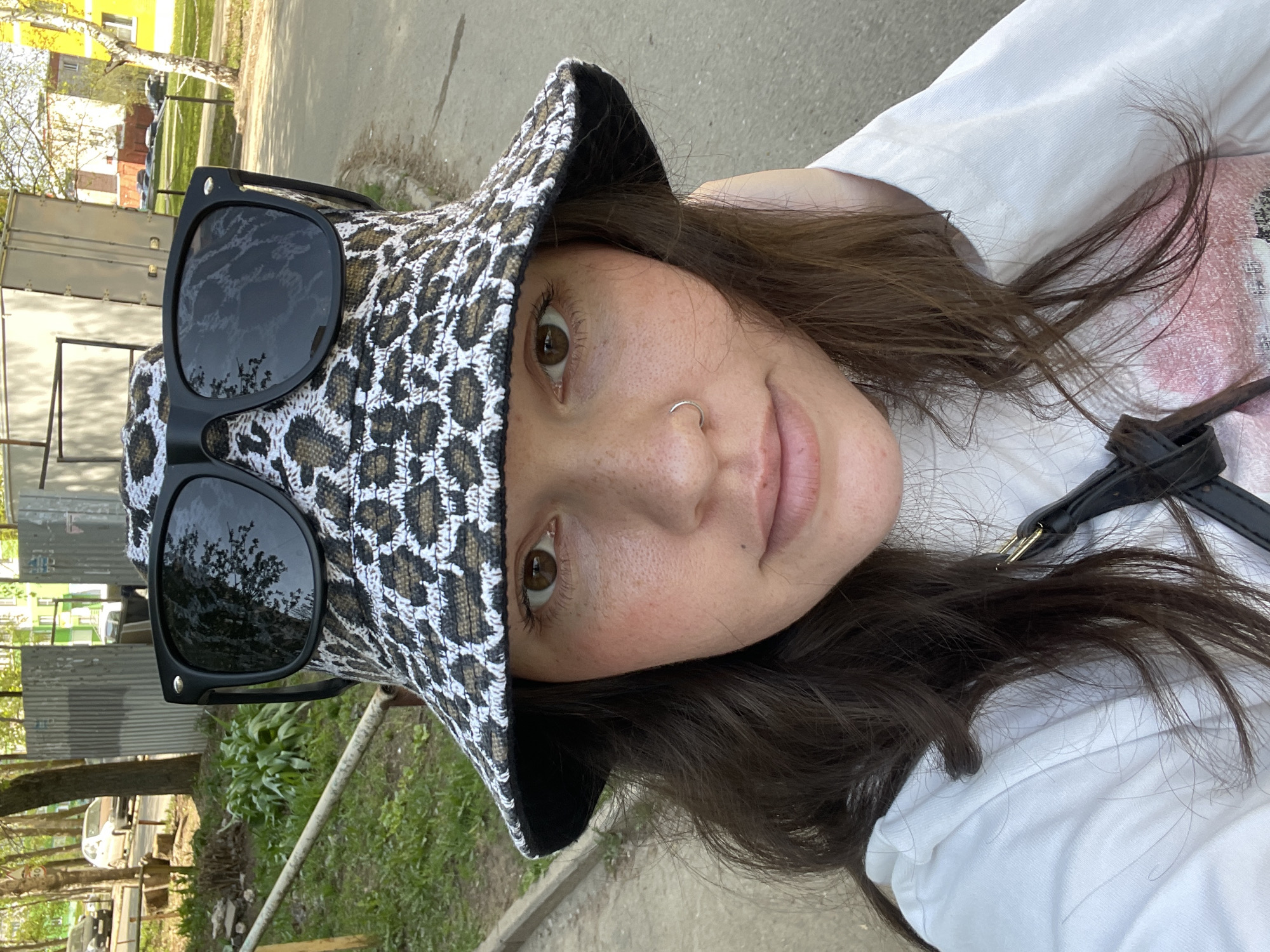 A woman is posing for a selfie in an outdoor location that appears to be a park or green space in an urban area. She is wearing a white t-shirt, a bucket hat with a white and brown leopard print pattern, and black sunglasses perched on top of the hat. She has brown hair, fair skin with freckles, and a nose ring. The surroundings suggest a residential or suburban area with buildings and trees visible in the background. A road or pathway is visible in the foreground, alongside a grassy area with vegetation. Based on background elements, it's potentially in Izhevsk, Russia, but requires more context for certainty.