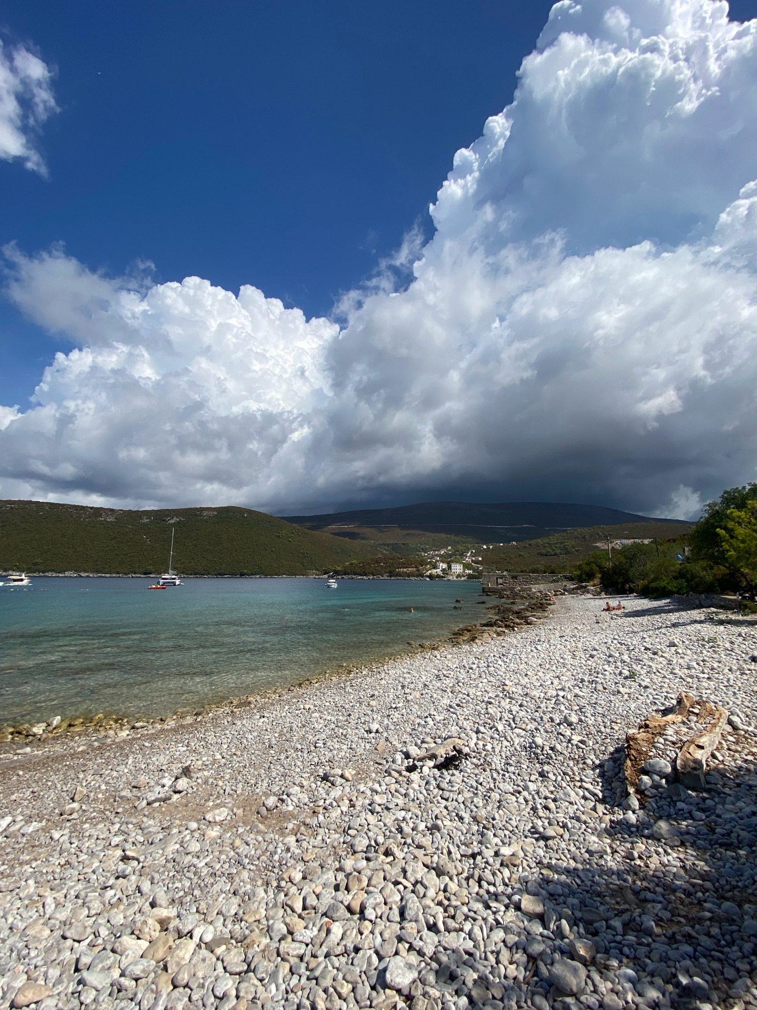 This image depicts a picturesque coastal scene in Luštica, Montenegro, under a dramatic sky. The foreground is dominated by a pebble beach, a natural expanse of smooth, grey and white stones. A large piece of driftwood rests on the beach, adding a rustic element. The water's edge meets the pebble shore, with clear, turquoise water revealing the submerged stones.
In the midground, the tranquil bay is dotted with several white sailboats and a small red dinghy, anchored in the calm waters. Beyond the bay, a lush, green hillside rises, covered in dense vegetation and trees. Small white buildings are scattered along the base of the hill, suggesting a small settlement.
The sky above is a dynamic contrast of bright blue and heavy, dark clouds. Towering white cumulus clouds dominate the upper portion of the frame, casting shadows and creating a sense of depth and movement. In the distance, the sky darkens significantly, hinting at approaching weather or a distant storm. A few small figures can be seen on the beach in the middle distance, appearing as red and orange specks on the pebbles. The overall impression is one of natural beauty, with a hint of atmospheric drama.