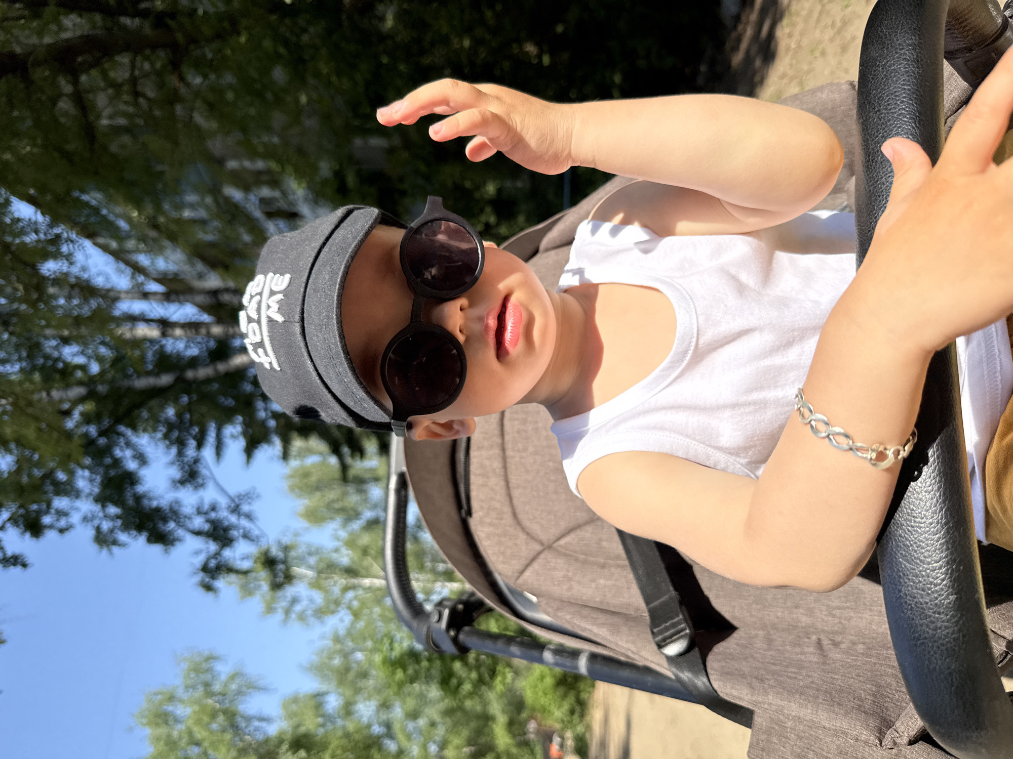 A young child is in a stroller. The child is wearing a white tank top, a black baseball cap, and black sunglasses. The child has a silver bracelet on their left wrist. The child is holding onto the stroller's handlebar. The background includes trees and blue sky. The image was taken in Rusanovo, Russia.