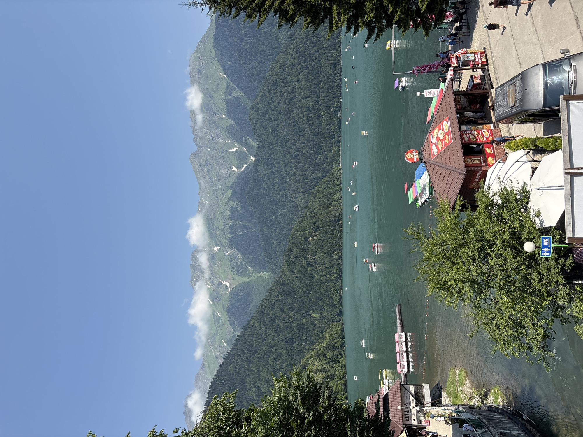 The image depicts a picturesque lake surrounded by lush green mountains under a clear blue sky. The water is a tranquil turquoise, dotted with numerous small boats, including pedal boats and motorboats. Along the shore, there are several buildings, including a cafe or restaurant with outdoor seating, a small shop selling food, and a dock with more boats. People are visible walking along the shore and near the buildings. The mountains in the background are covered in dense forest, with some patches of snow visible on the higher peaks, suggesting a high altitude location. The overall scene conveys a sense of leisure and natural beauty. The location is identified as Lake Ritsa, Abkhazia.