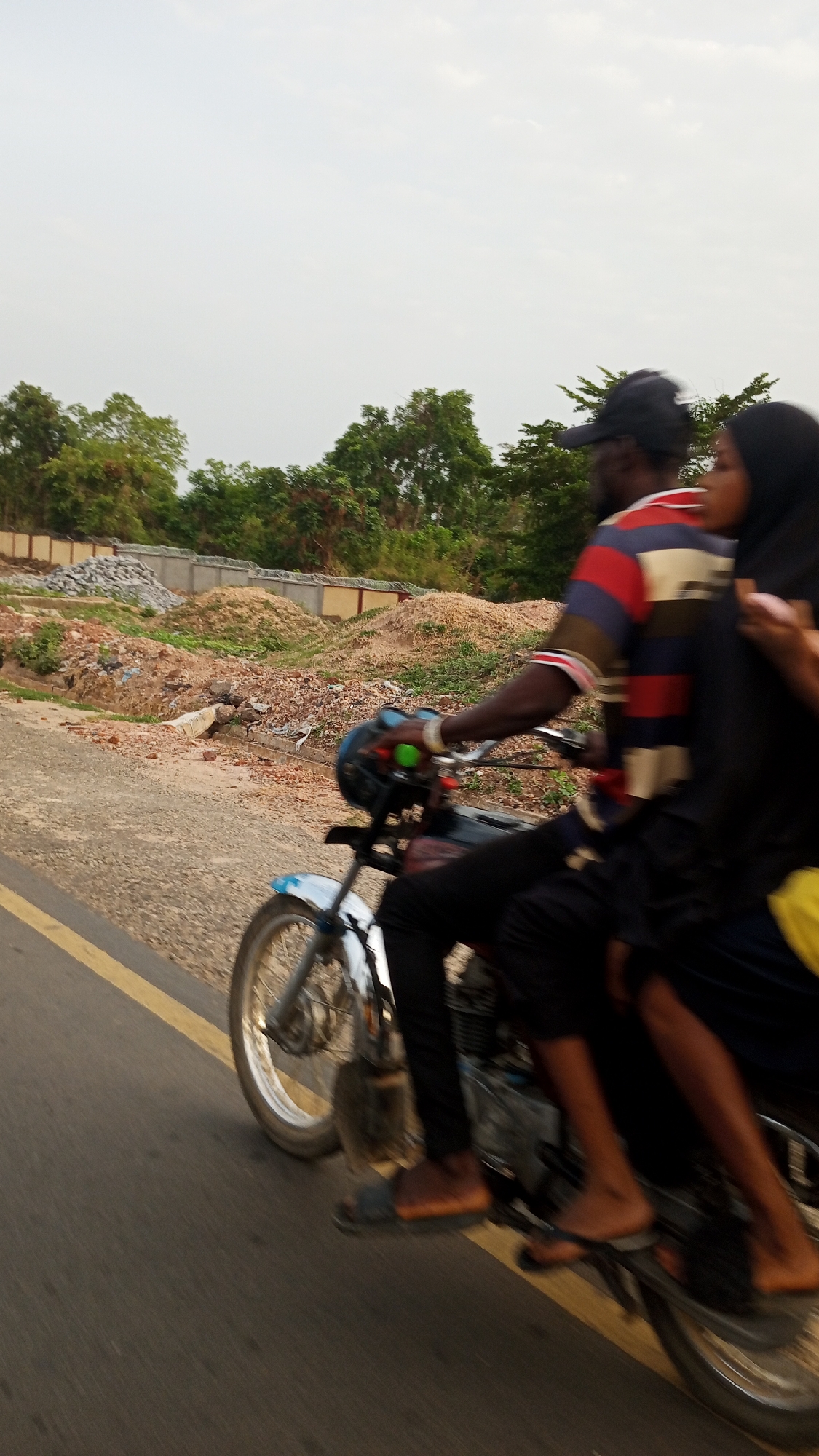 A blurred image shows two individuals riding a motorcycle on an asphalt road marked with a yellow line. The rider, wearing a striped shirt and a baseball cap, is operating the motorcycle. The passenger, wearing what appears to be a hijab, is seated behind the rider. Both are wearing sandals.
The background features trees, some construction materials, and structures. The location appears to be Zango-ndaji, Nigeria. The movement of the motorcycle is evident, as the surrounding environment is blurred.