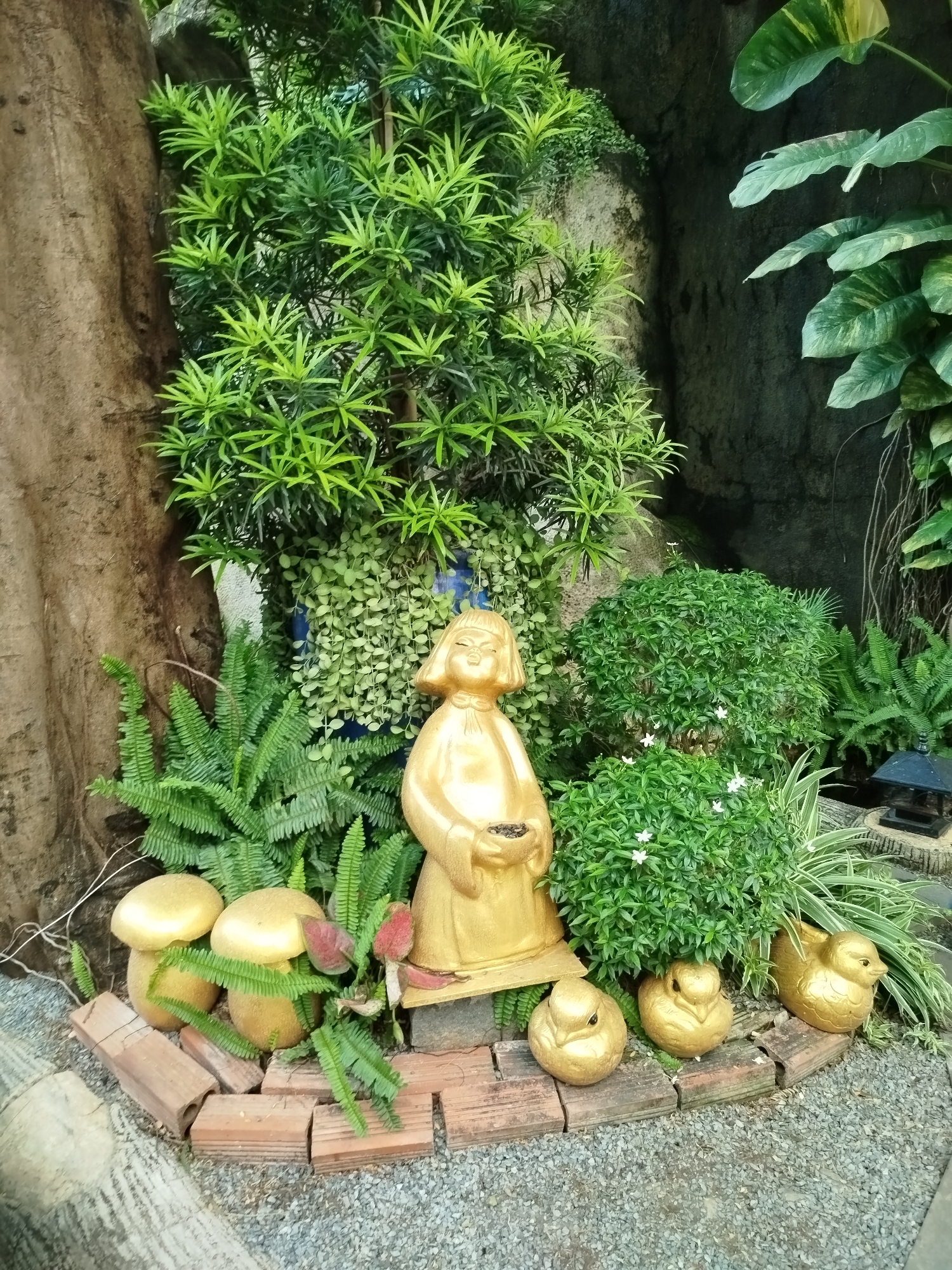 The image depicts a small garden scene, likely located in Ho Chi Minh City, Vietnam. A golden statue of a person, possibly a deity or a folkloric figure, stands prominently in the center. Around the statue, there are several golden bird figurines, arranged in a row along a short brick border. Behind the statue are various plants, including ferns, small-leaved shrubs with tiny white flowers, and a taller shrub with needle-like leaves. To the left are golden mushroom ornaments. The background consists of a rock-like formation and some other greenery. The ground is covered with small gravel.