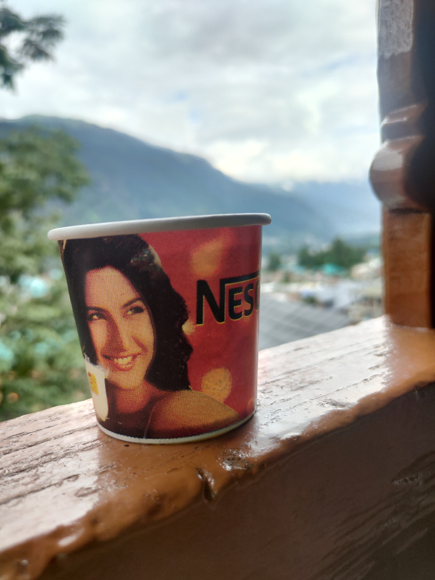 An outdoor, medium shot captures a disposable coffee cup placed on a wooden ledge. The cup, featuring a smiling woman's face and the "Nes" logo, is positioned in the foreground, slightly to the left of center. The background is a blurred landscape of mountains and a cloudy sky. The wooden ledge is textured and appears to be part of a railing or balcony. The scene suggests a relaxed moment with a view, possibly in a scenic location. The overall atmosphere is serene and natural, with a hint of human presence through the coffee cup.