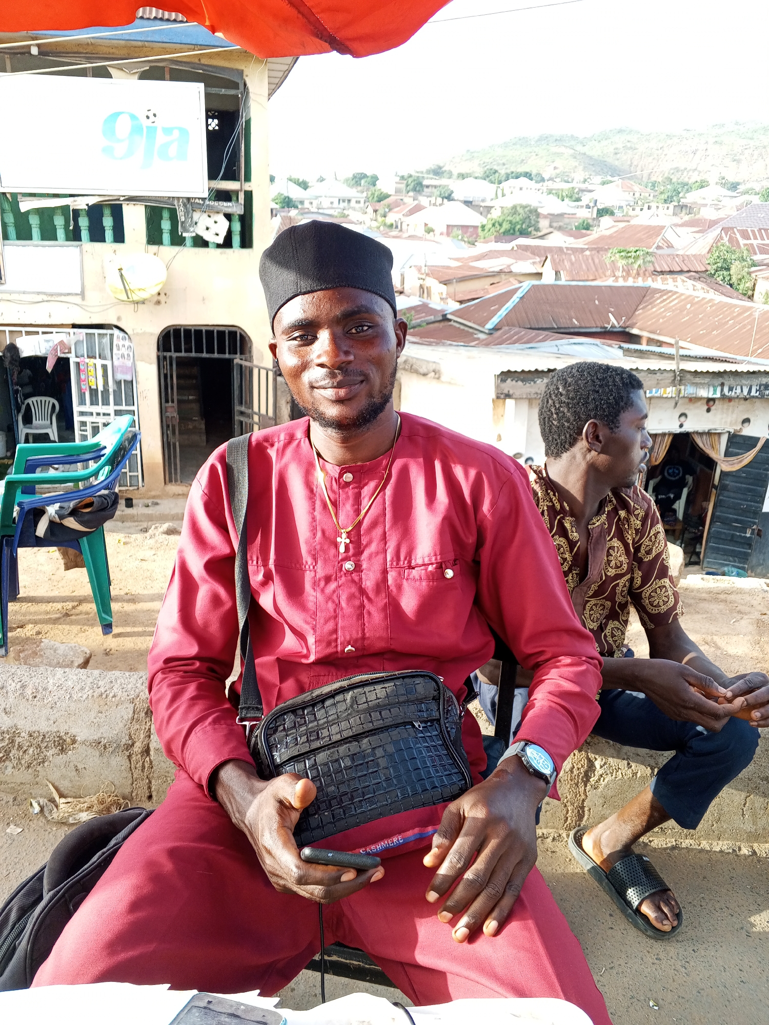 Two men are seated outside in what appears to be the city of Gwong, Nigeria. The foreground features a man wearing a maroon shirt and pants, a black cap, and a gold cross necklace. He is carrying a black shoulder bag and holding a phone. Seated behind him to the right is another man wearing a patterned shirt and dark pants and slides. The background shows buildings with corrugated metal roofs and a hillside with sparse vegetation. A blue and green plastic chair sits to the left, and a sign with the text "9ja" is visible above a doorway.