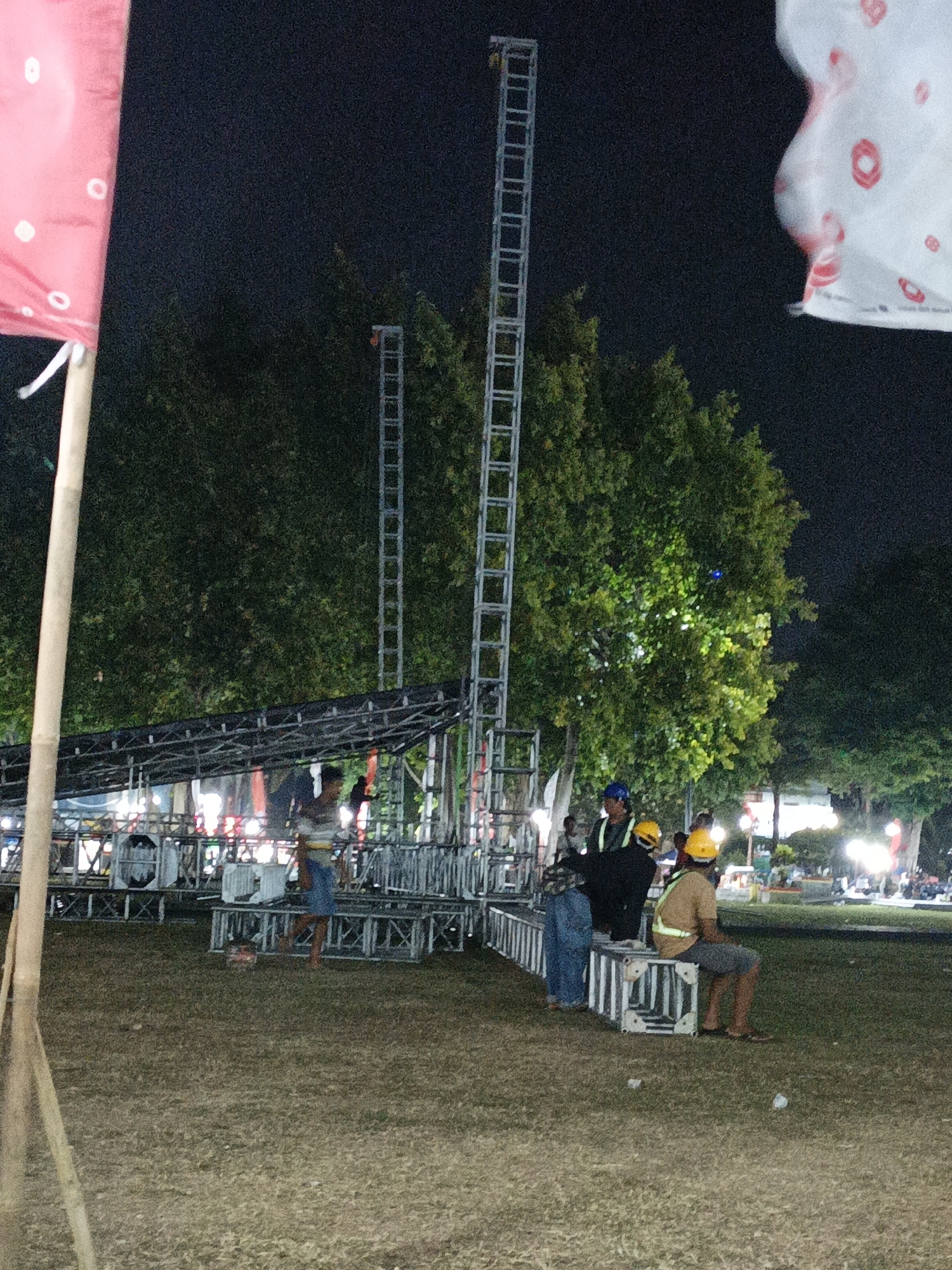 In Klaten, Indonesia, at night, a group of people are gathered around a stage setup. Two tall, ladder-like structures, likely for lighting or sound equipment, are visible behind the stage. The stage itself is constructed from metal scaffolding and appears to be in the process of being set up or taken down. Several individuals are present, some wearing hard hats and high-visibility vests, suggesting they are workers. One person is standing on the stage, while others are seated or standing near it. In the foreground, a tall wooden pole with a red and white flag is partially visible. The ground is covered in dry grass, and trees form a dark backdrop against the night sky. Lights from the stage area and in the distance illuminate the scene.